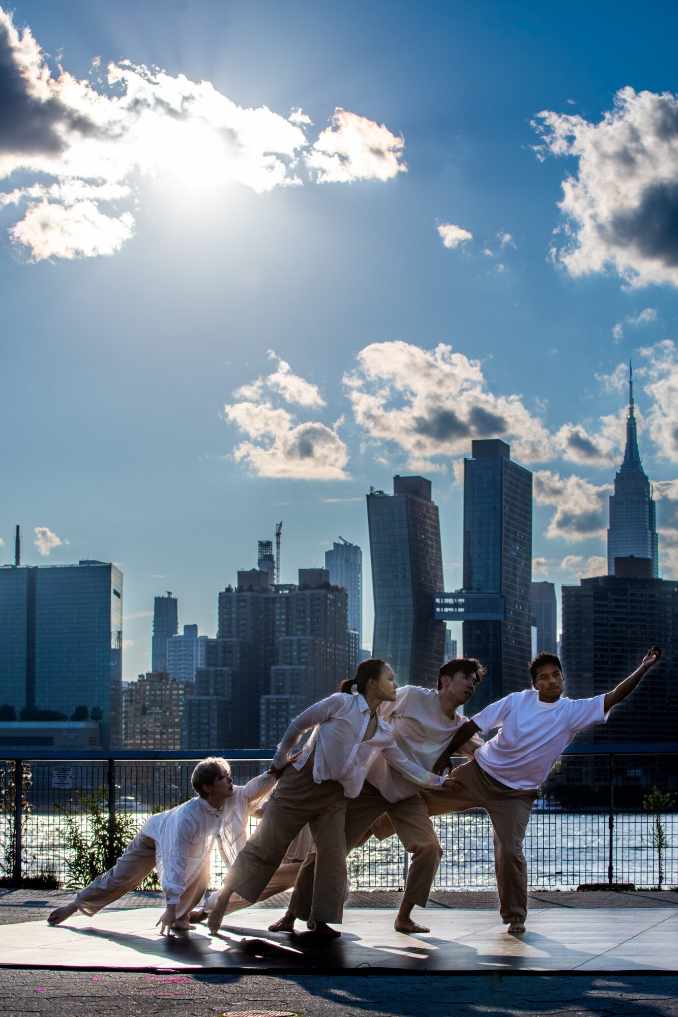 People dance on a waterfront promenade with a city skyline and bright clouds behind them.