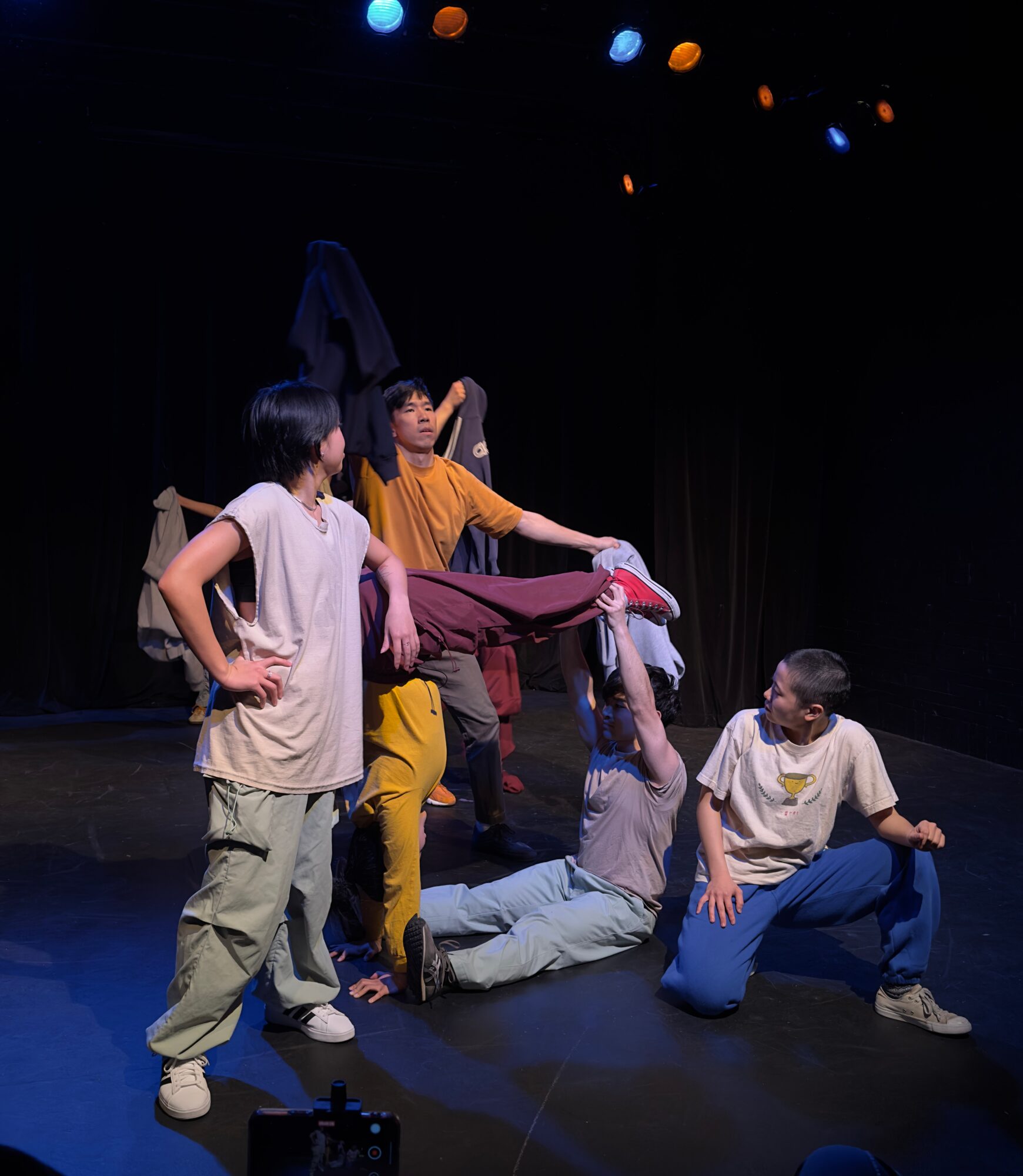 Several people pose on a dark stage beneath colored lights, with one person seated on the floor and others gathered around.