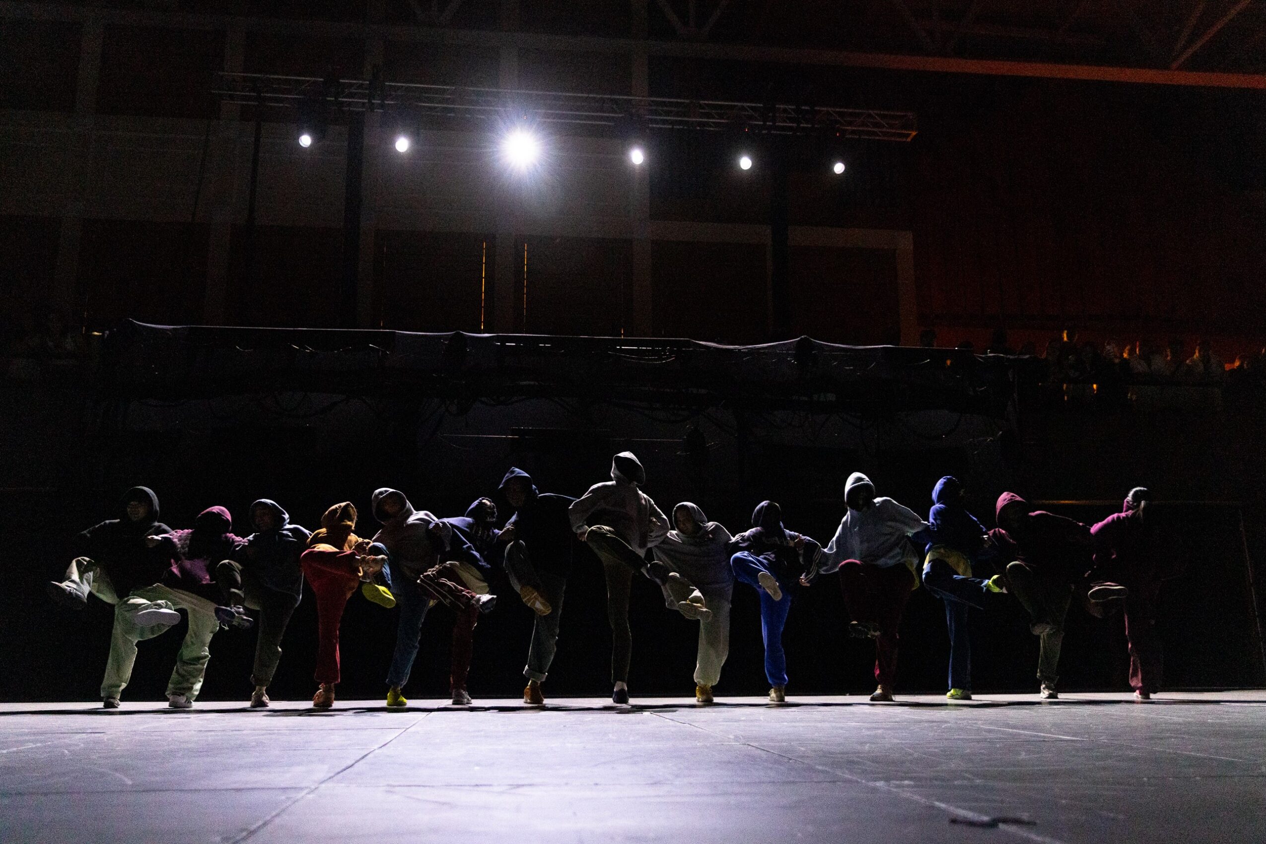 A line of people performs on a dimly lit indoor stage beneath overhead lights.