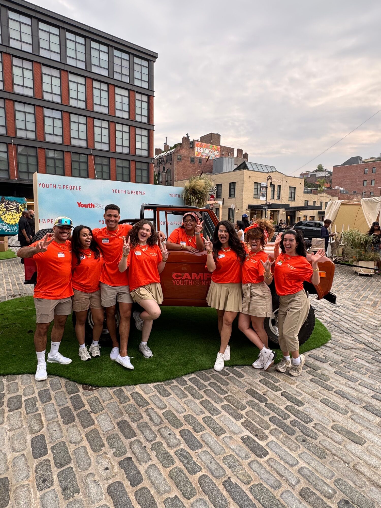 A group in orange shirts poses together on a small patch of grass beside a cobblestone street.