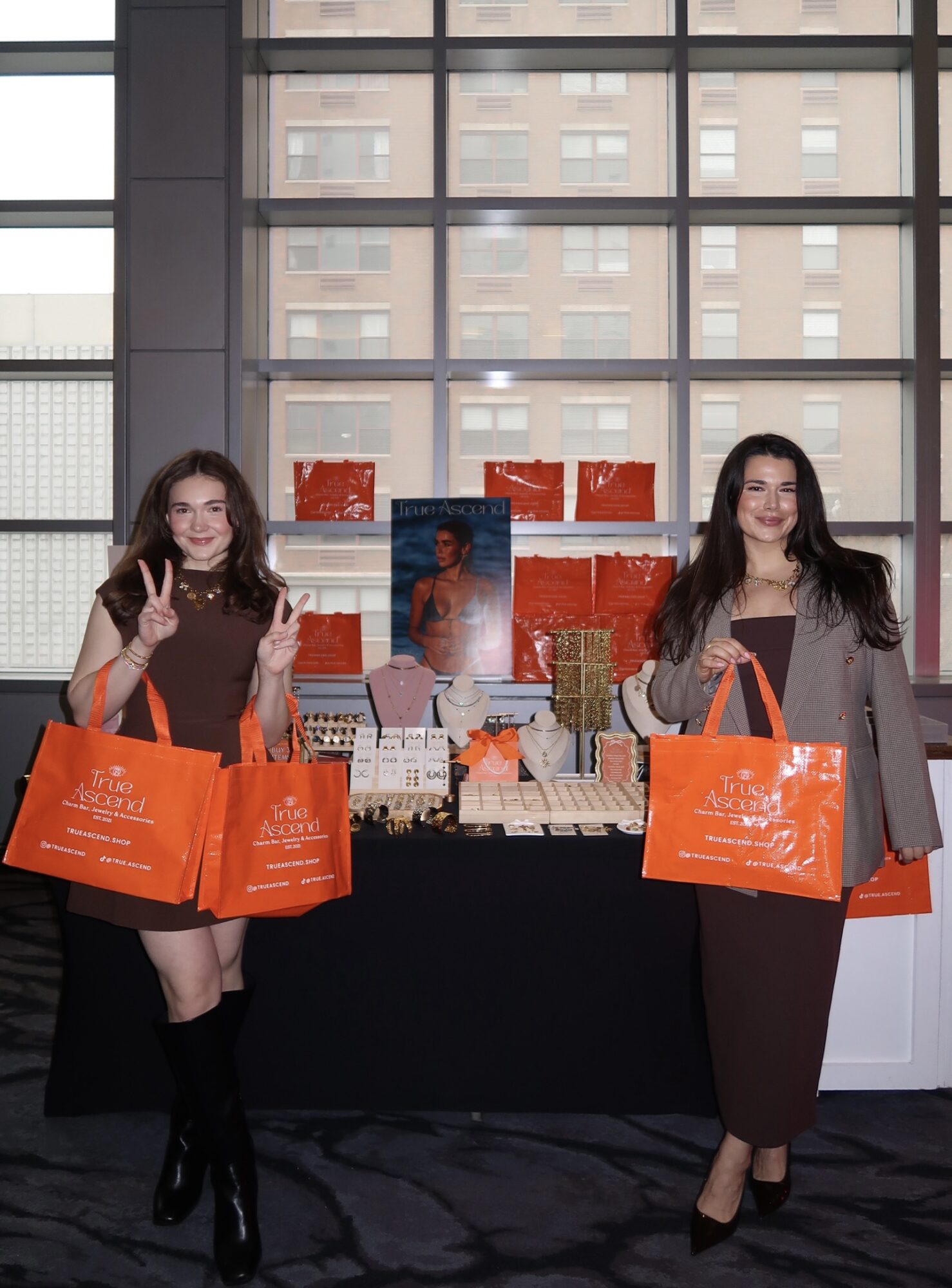 Two people stand behind a display table with orange bags, books, and red signs.