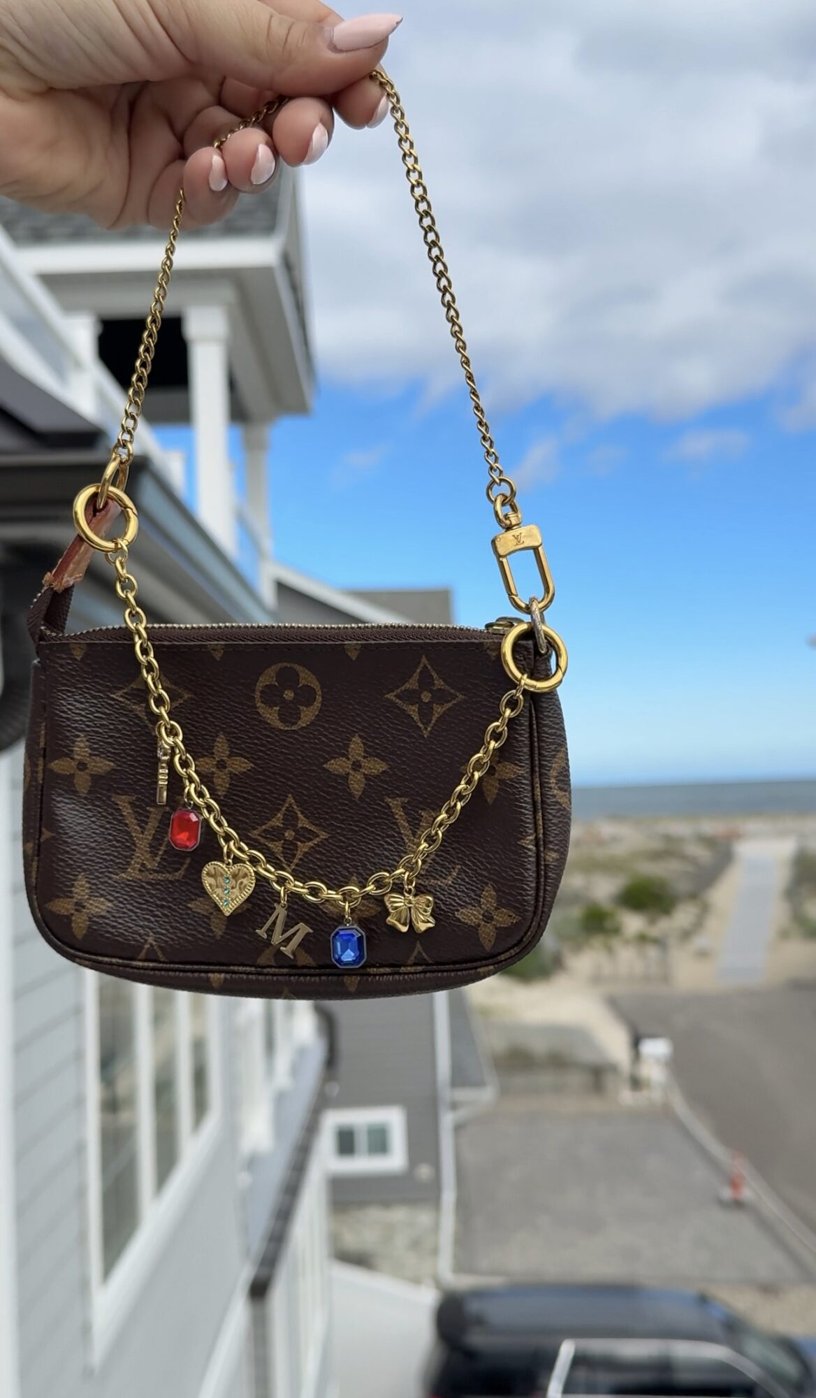 A hand holds a brown patterned handbag by its gold chain against a coastal view from a balcony.