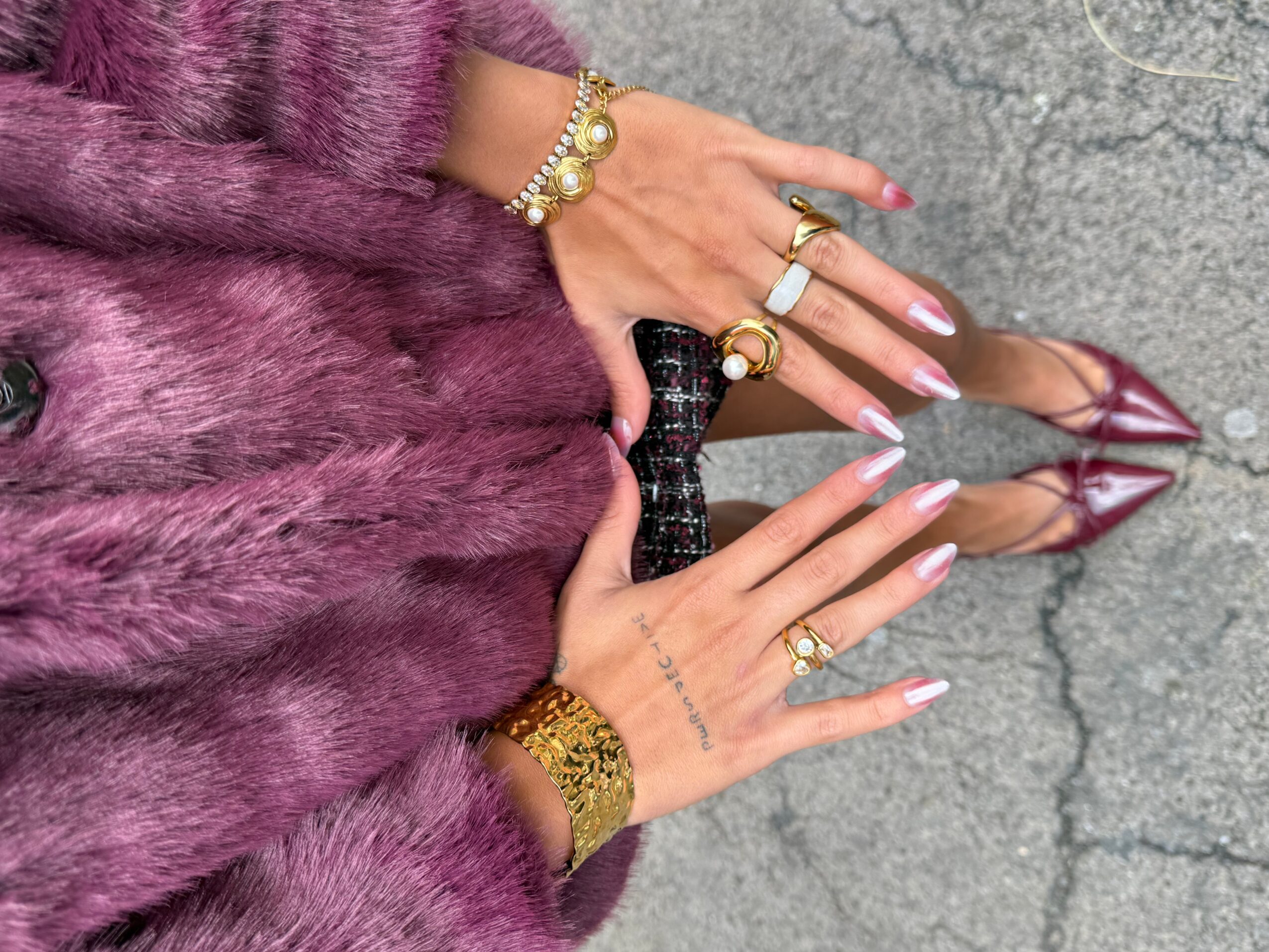 Hands with long manicured nails and jewelry hold a black purse over a purple fur coat and cracked pavement.