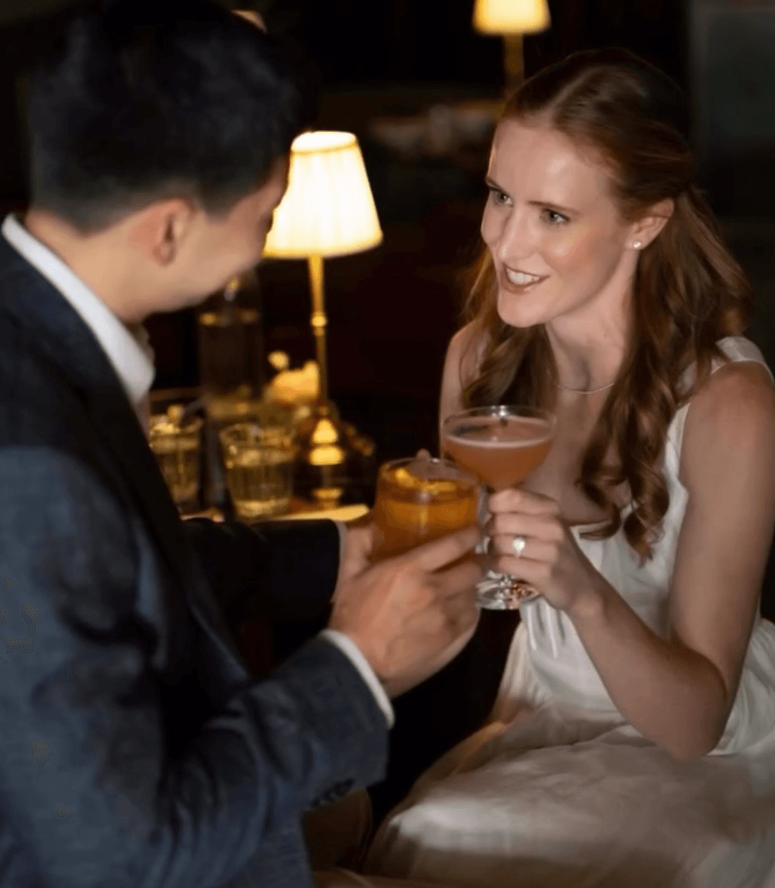 A seated couple clink drinks at a candlelit table; the woman holds a cocktail glass and the man holds a tumbler.