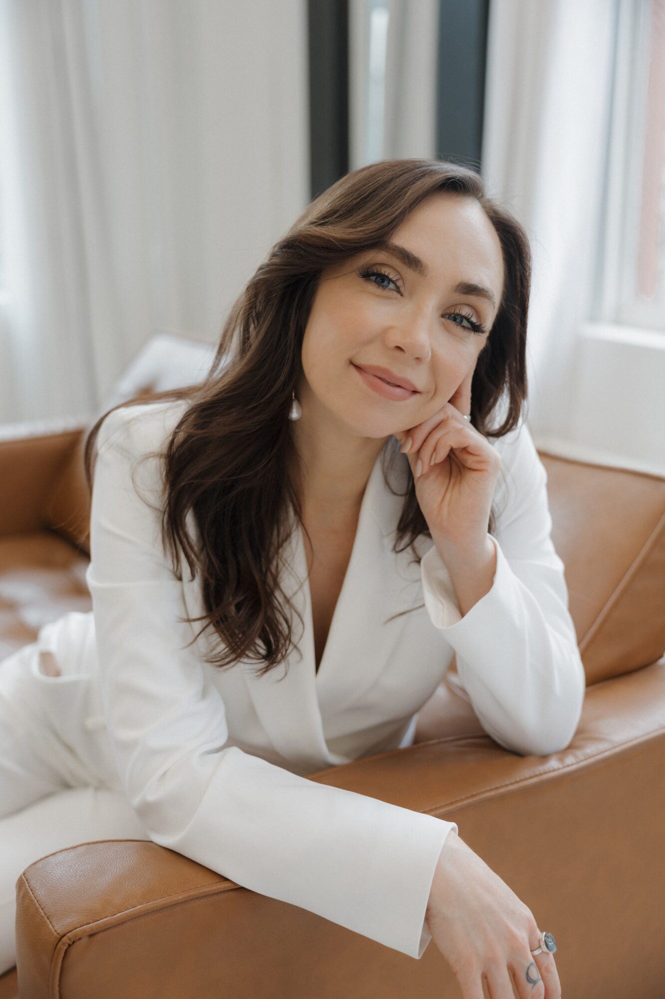A woman in a white suit sits on a tan leather chair, resting her chin on one hand.