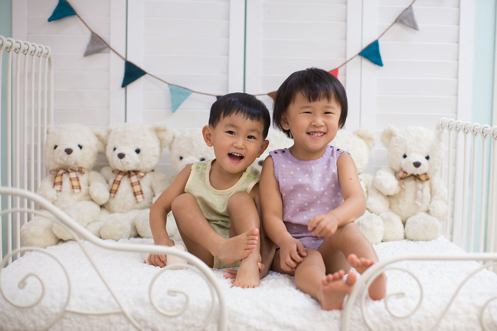 Two children sit together on a white bed among stuffed animals and triangular bunting.