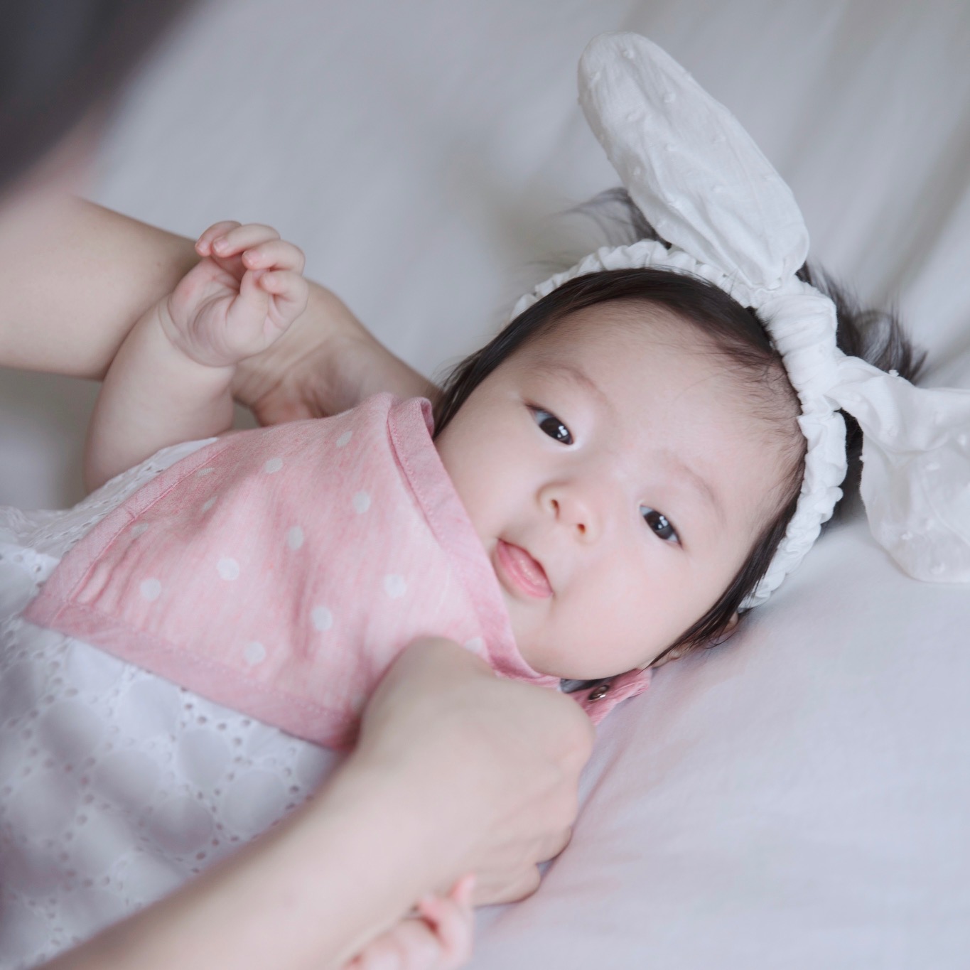 Baby lying on a white blanket, wearing a pink polka-dot outfit and a large white bow headband.