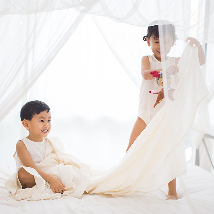 Two children play with white fabric beneath sheer curtains.