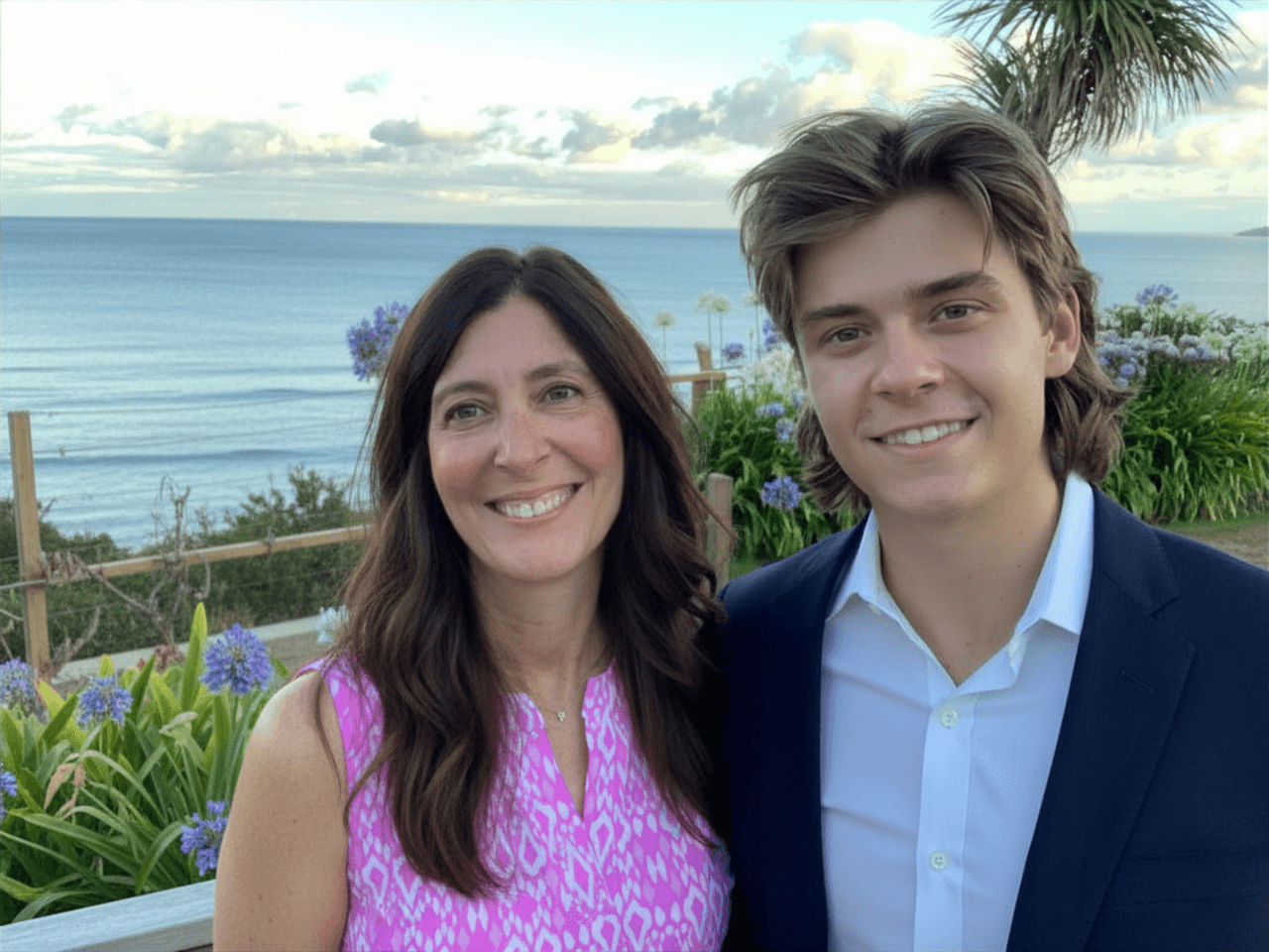 A woman and a young man pose by a garden with ocean water in the background.