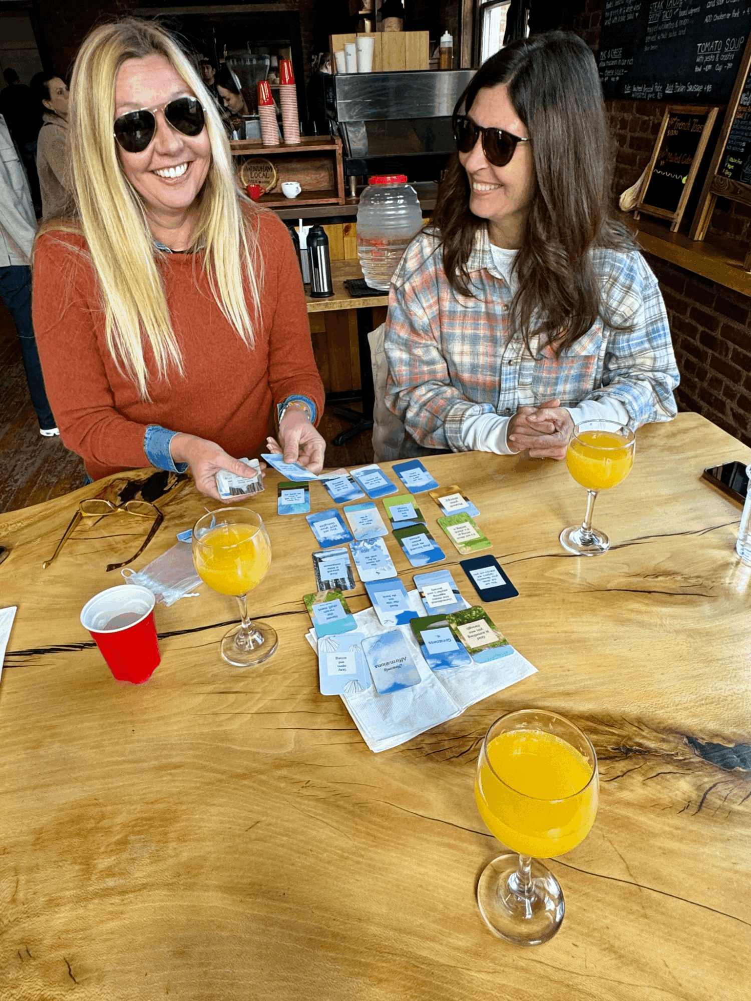 Two people wearing sunglasses sit at a wooden table with cards, drinks, and a red cup.