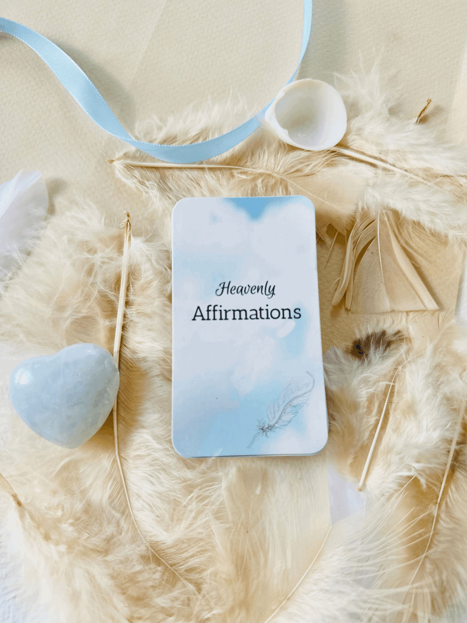 A blue affirmation card rests among white feathers, with a ribbon, small ornament, and heart-shaped stone nearby.