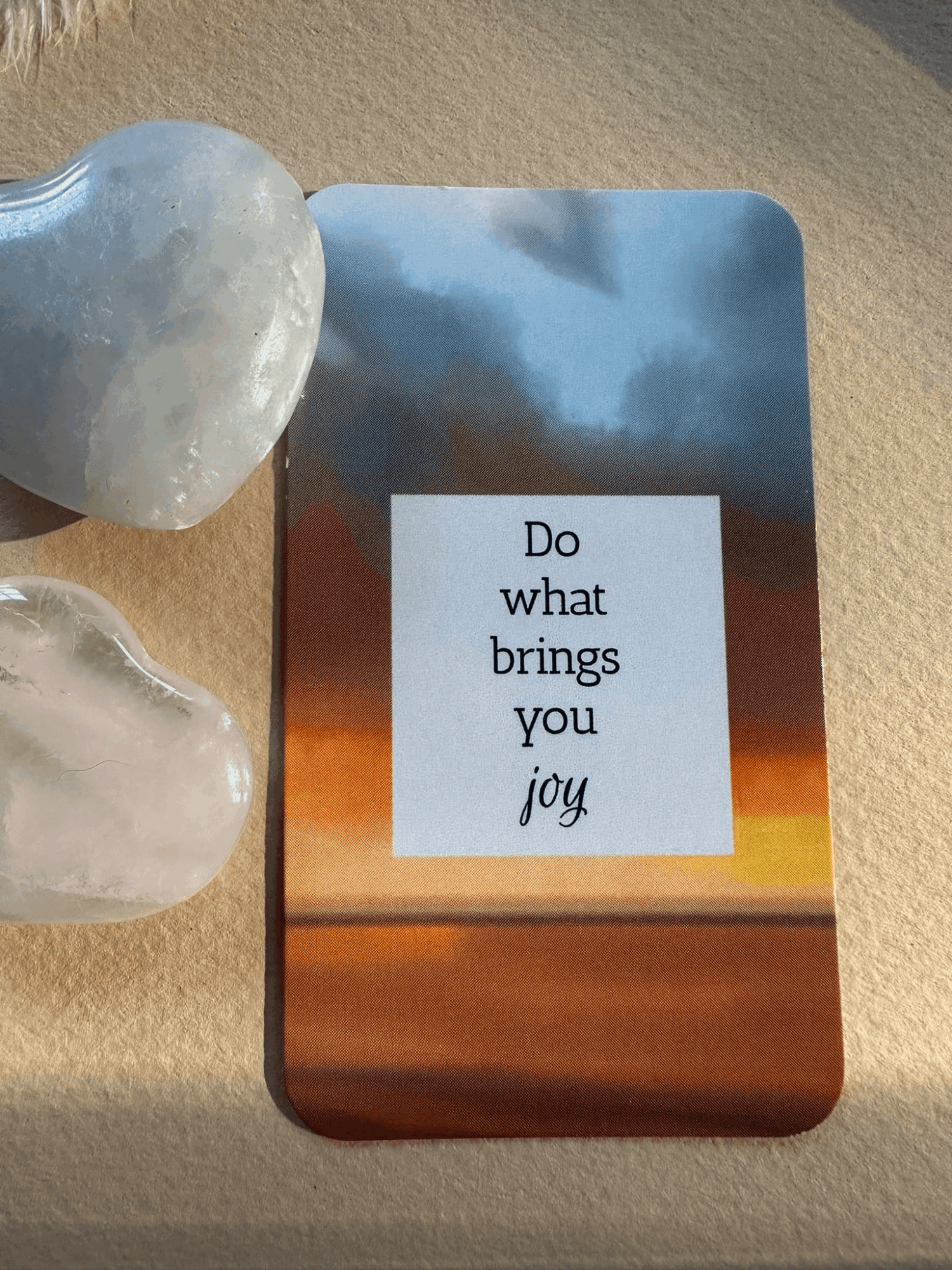 A card reading “Do what brings you joy” beside two translucent stones on a textured surface.