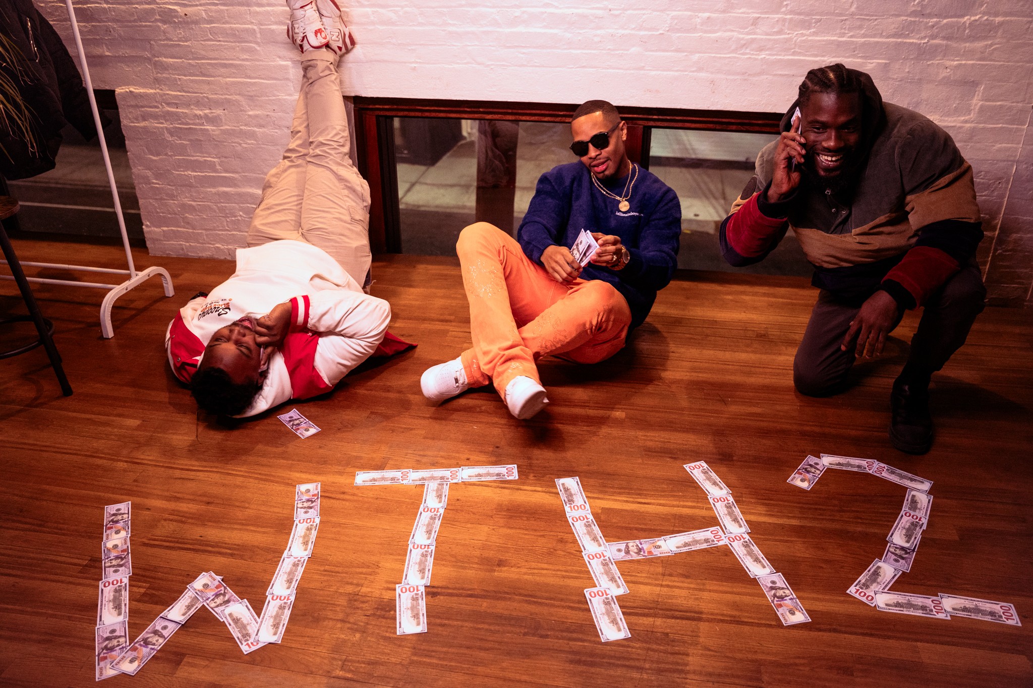 Two people sit on a wooden floor beside a person lying down; paper letters spelling “WITH” are arranged nearby.