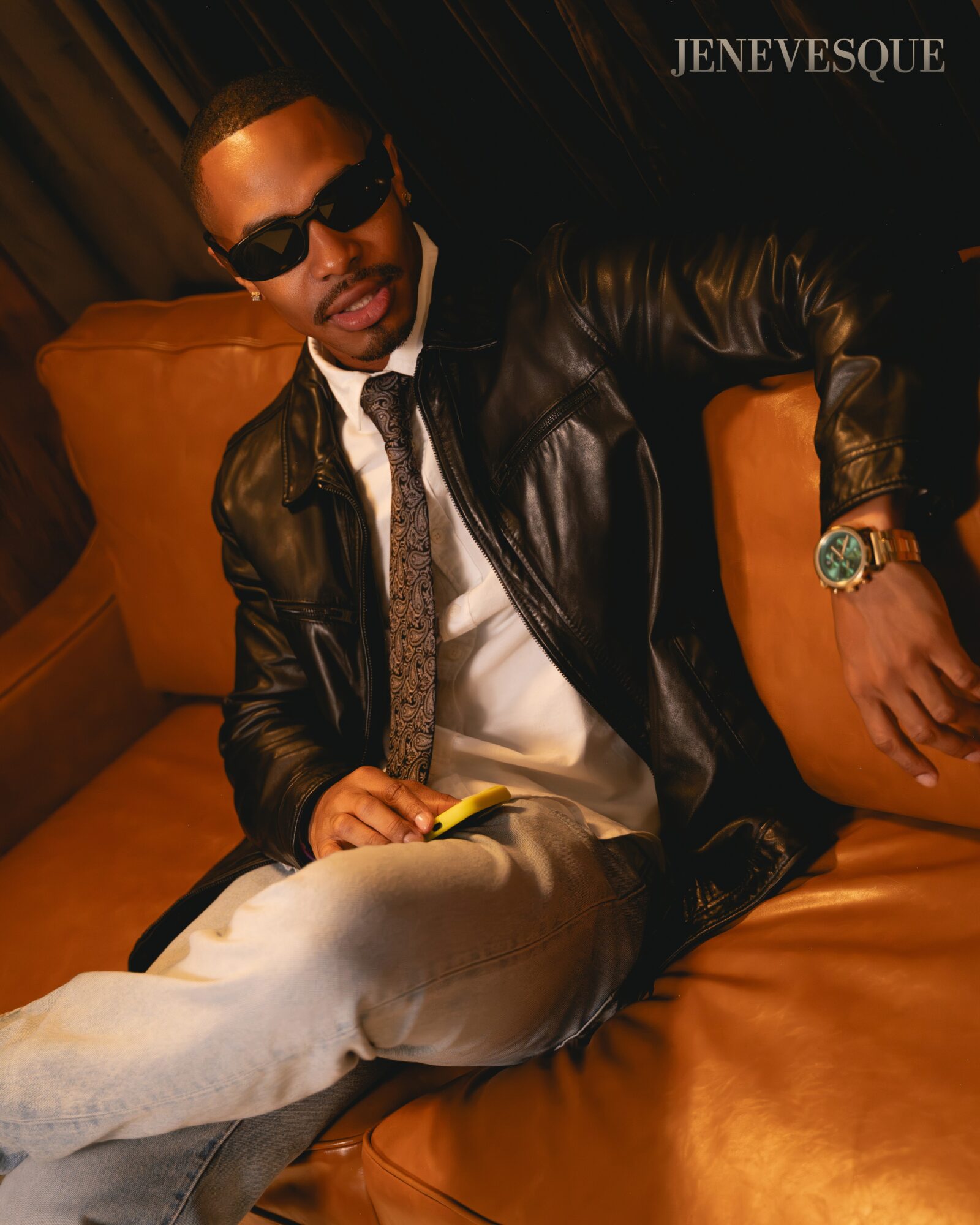 Person wearing sunglasses and a leather jacket seated on an orange leather couch.