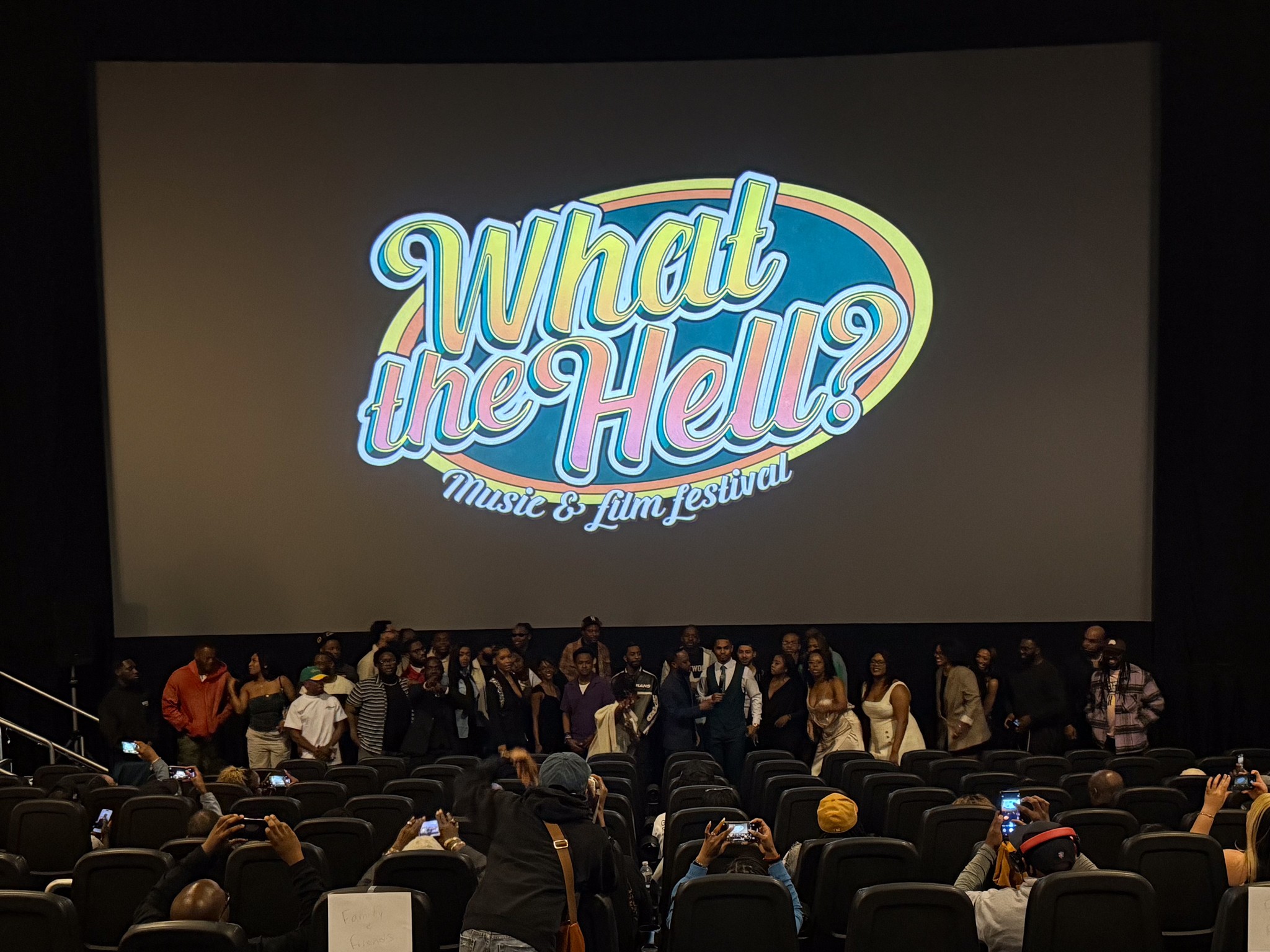 A cinema audience faces a large screen displaying the colorful title “What the Hell?,” with people standing near the front.