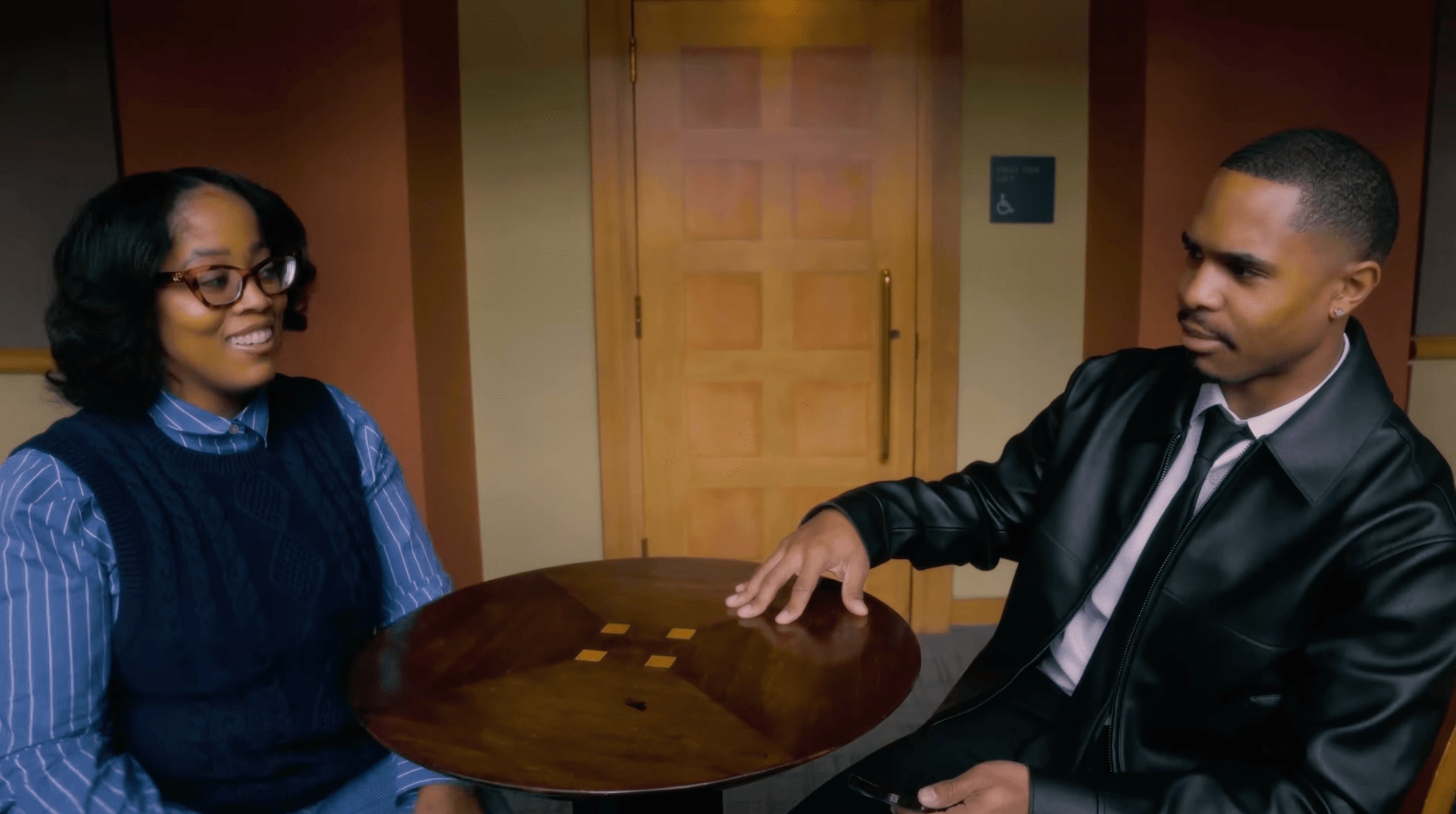 Two people sit across a round wooden table in a room with a closed door.
