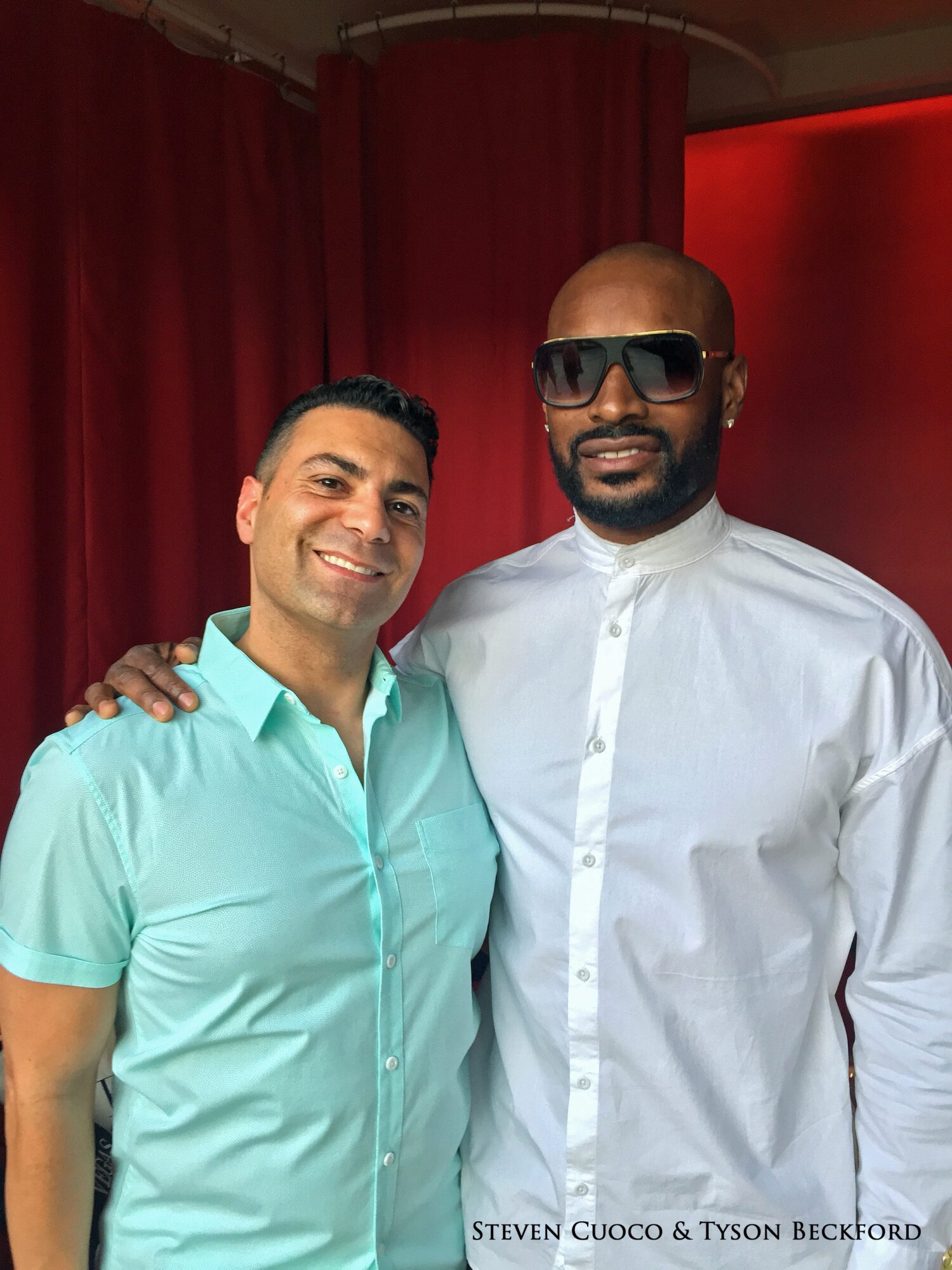 Two men stand together in front of red curtains; one wears a turquoise shirt, the other a white shirt and sunglasses.
