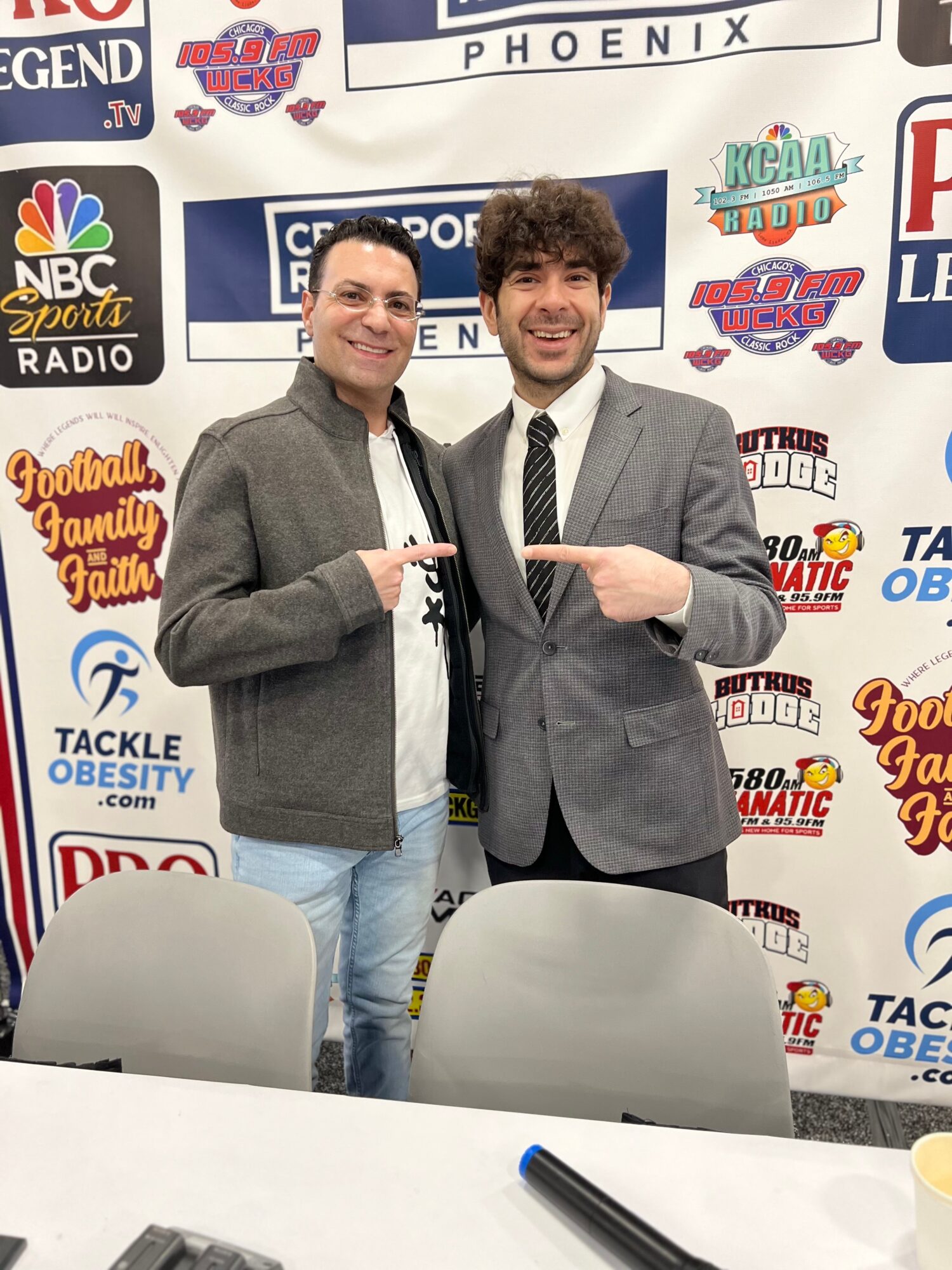 Two people stand in front of a sports-themed backdrop, pointing toward a sign between them.