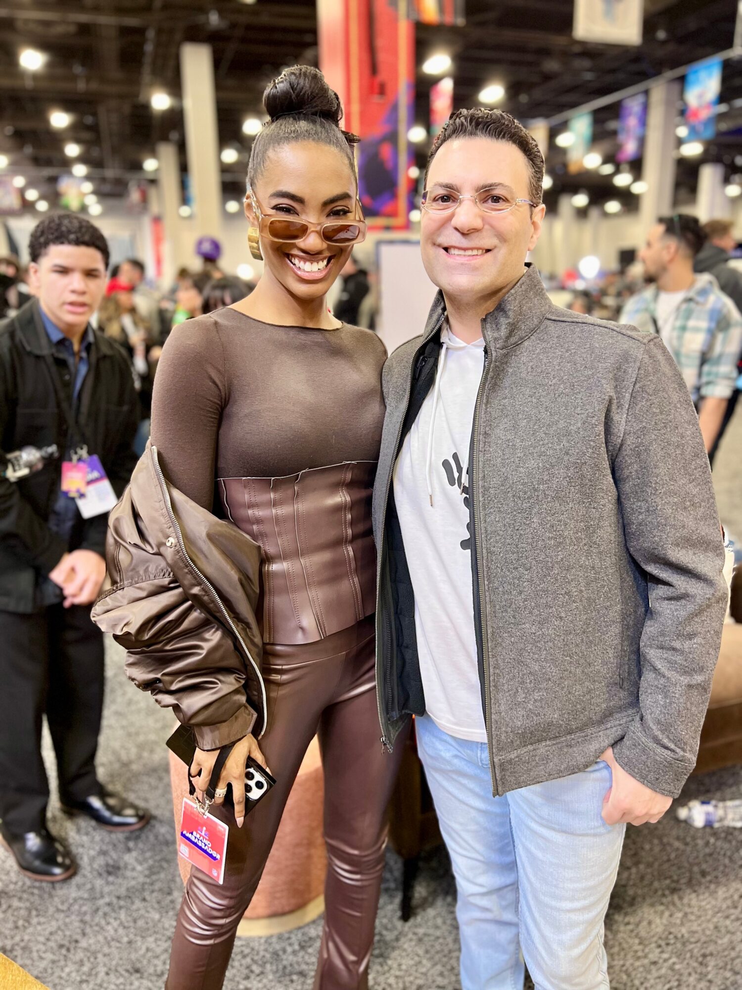 Two people pose together at a busy indoor event, with attendees and booths in the background.