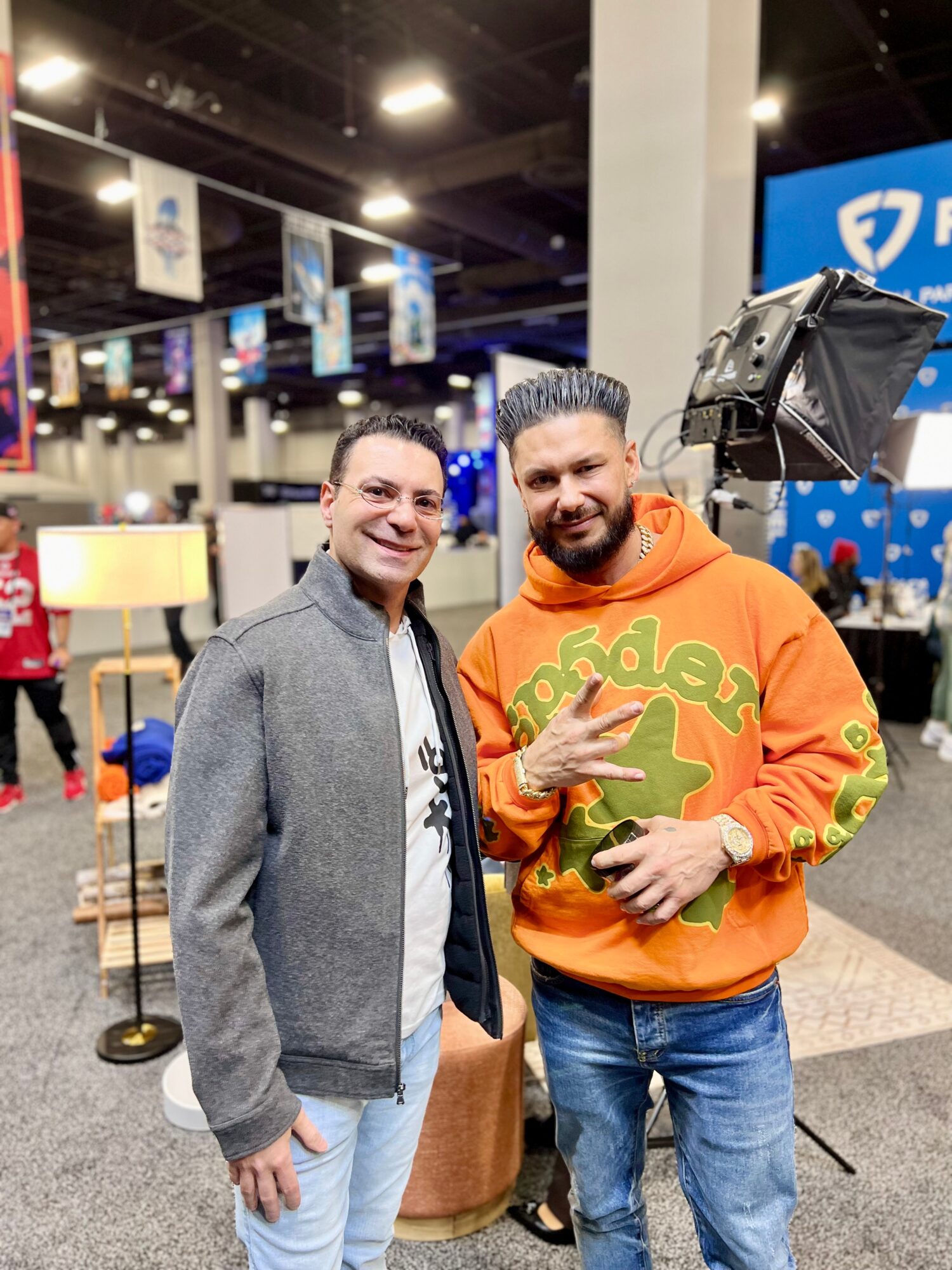 Two people pose together at an indoor event, one wearing a gray jacket and the other an orange sweatshirt.