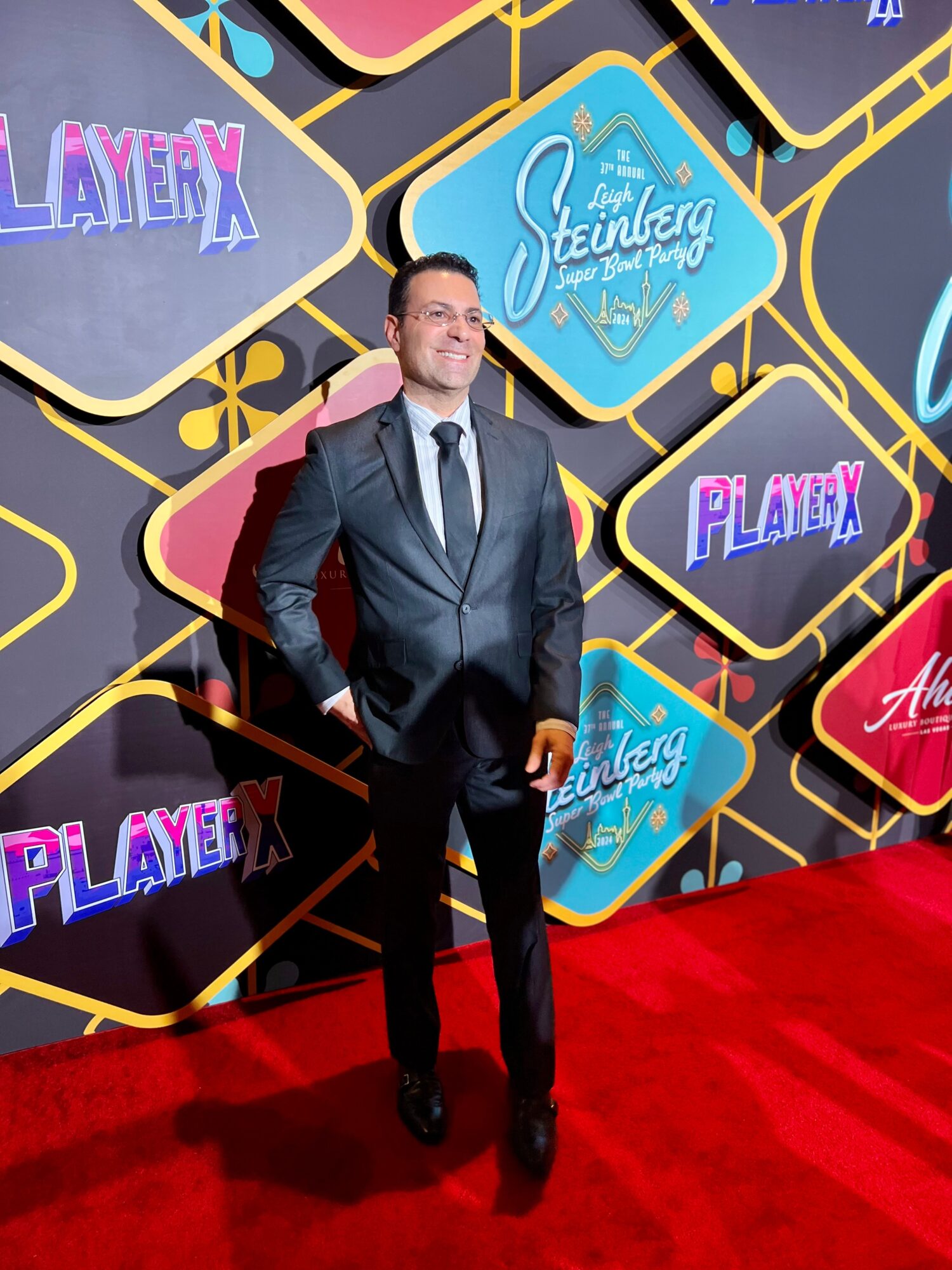 A person in a dark suit stands on a red carpet before a colorful event backdrop.