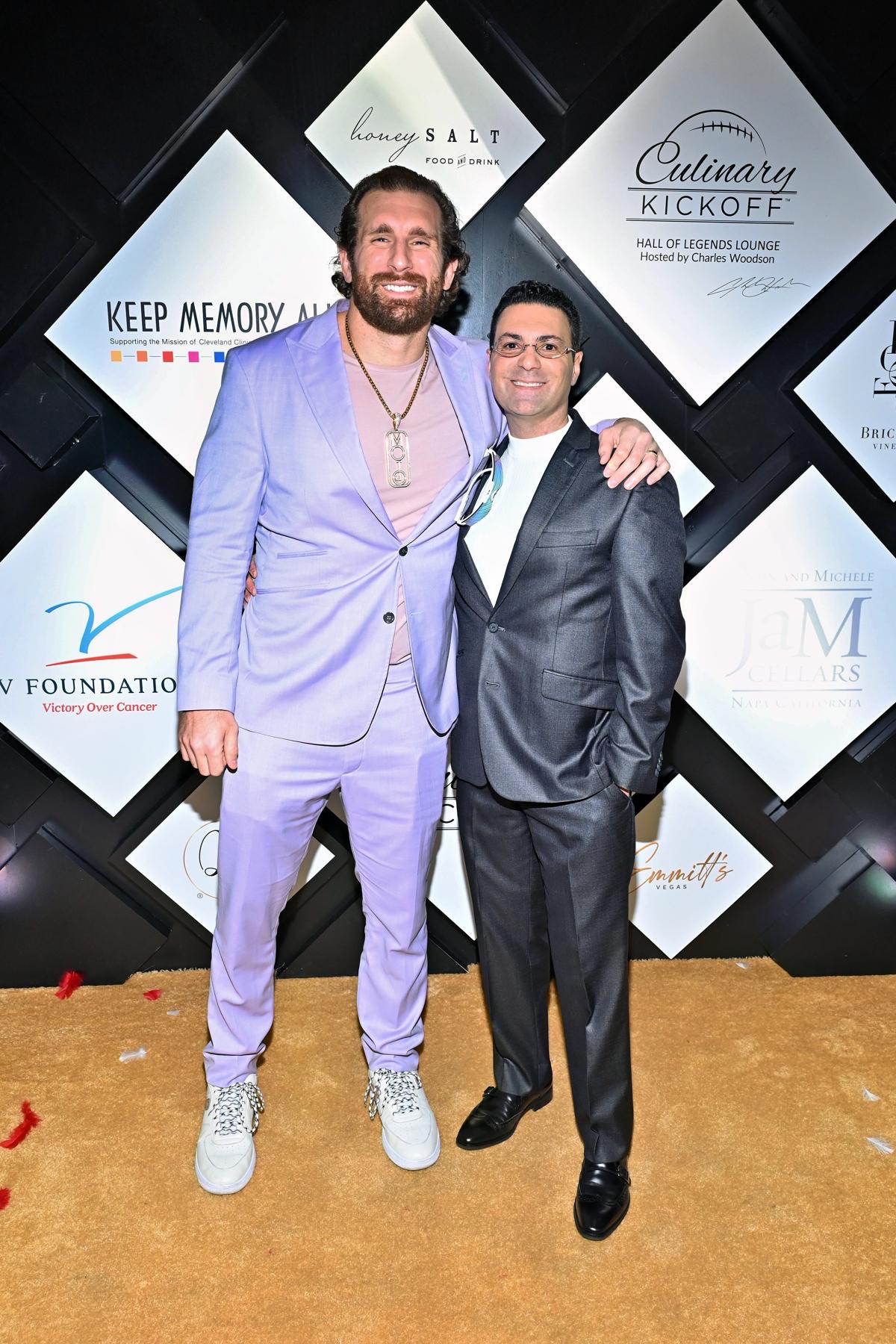 Two men stand together on a carpeted event floor in front of a sponsor-logo backdrop.