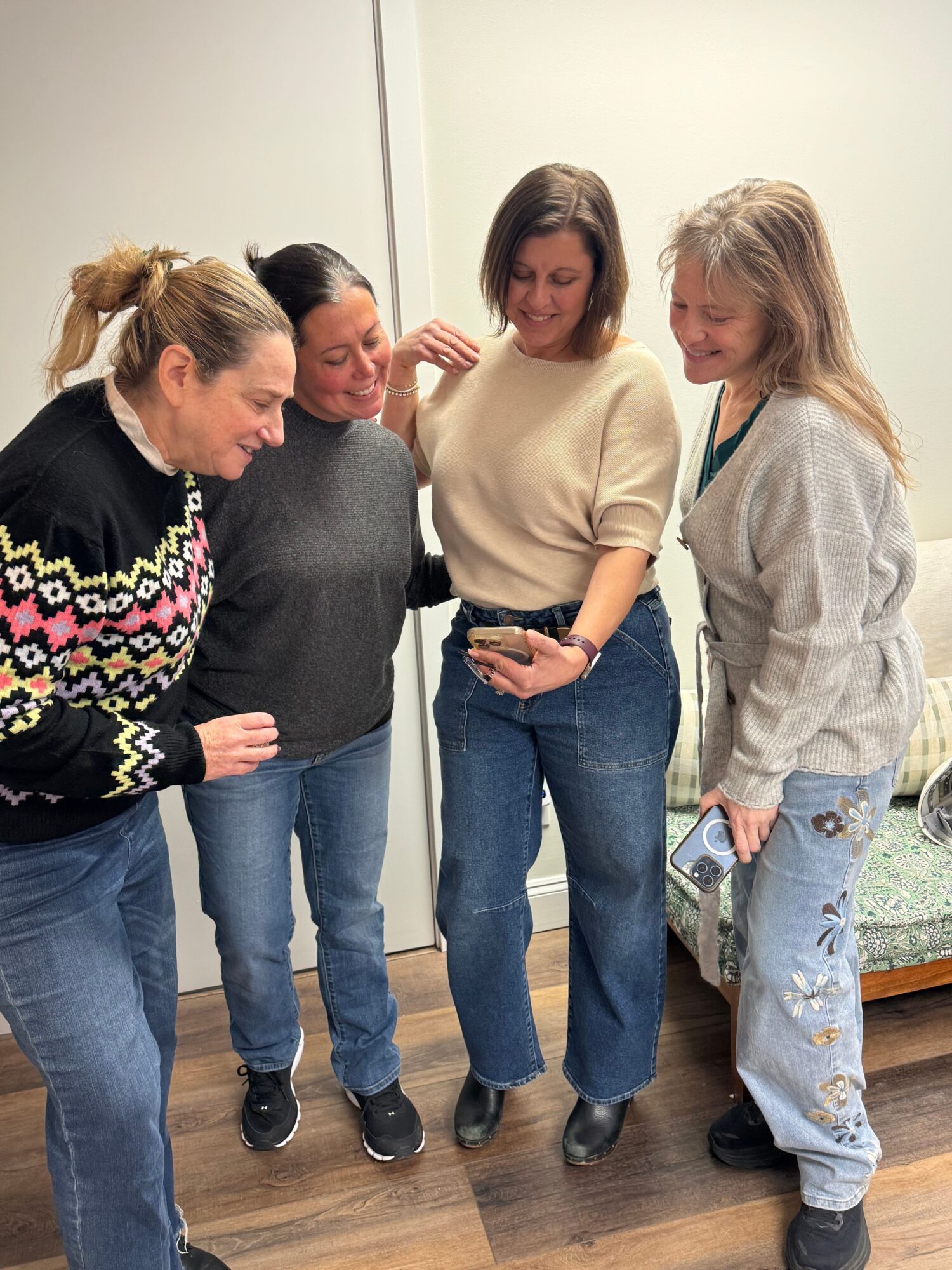 Four people stand together indoors, with a patterned sweater, jeans, and a phone visible.