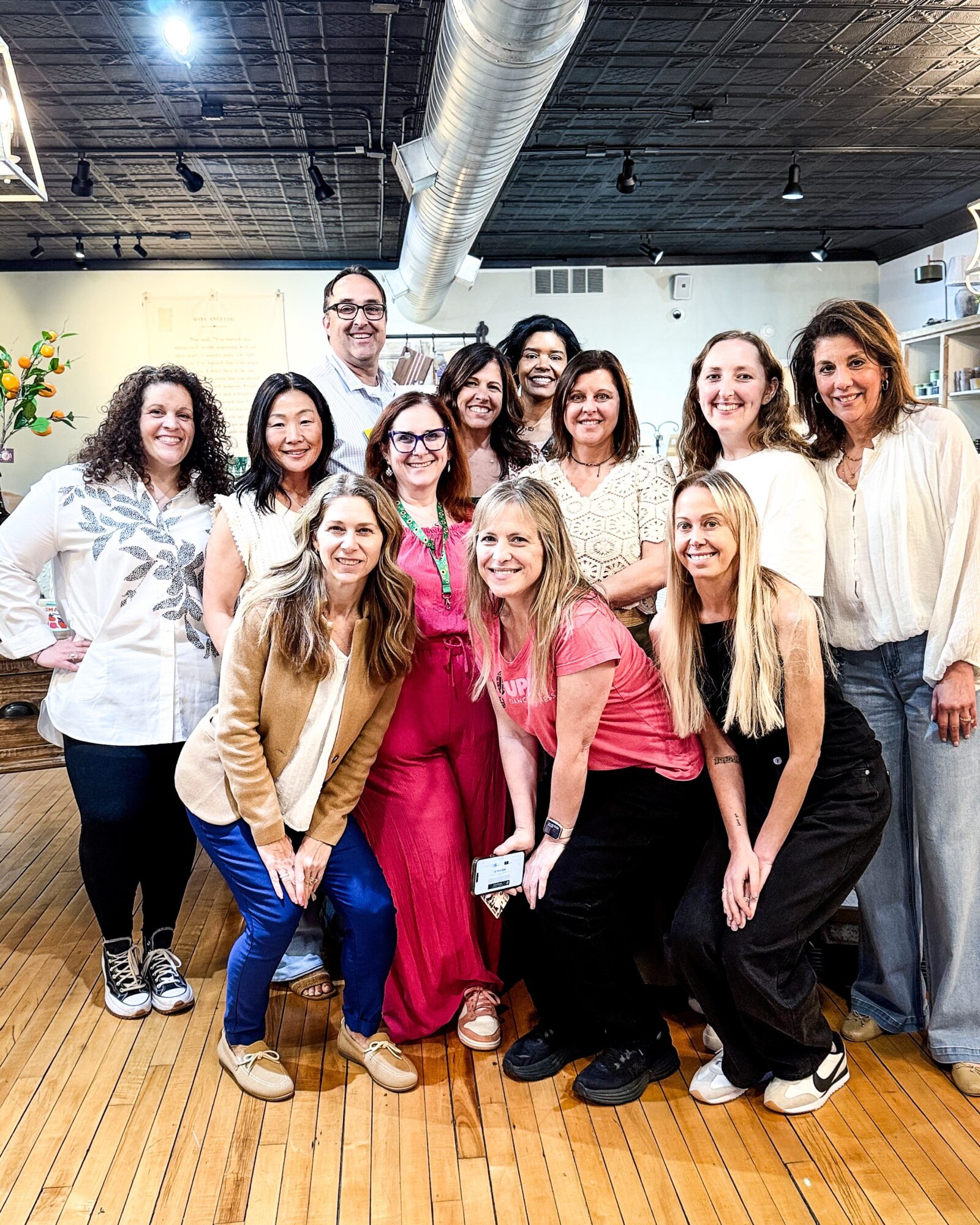 A group of people pose together in a bright room with wood flooring and exposed ceiling ducts.