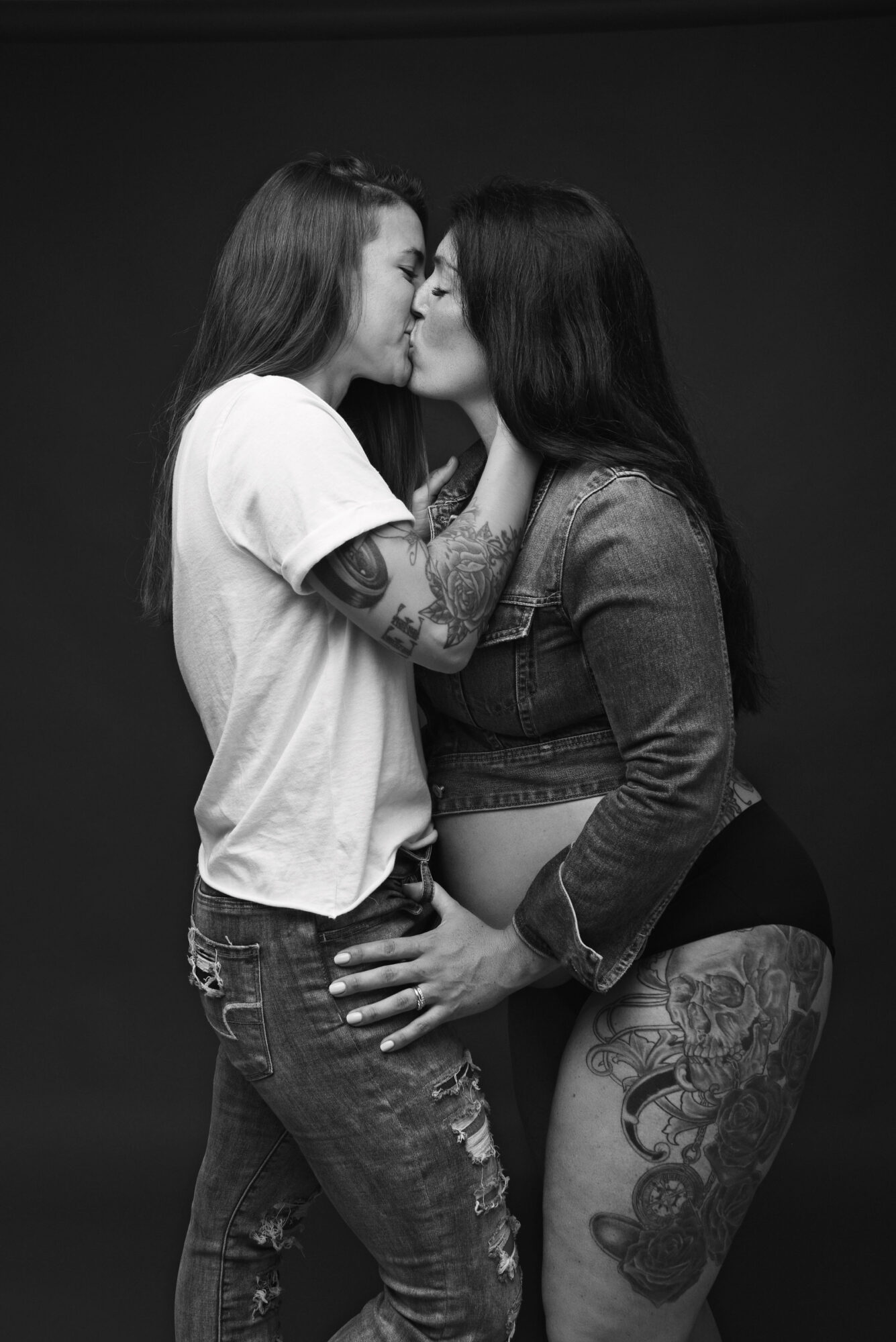 Two people kiss while standing against a dark background; both have visible tattoos.