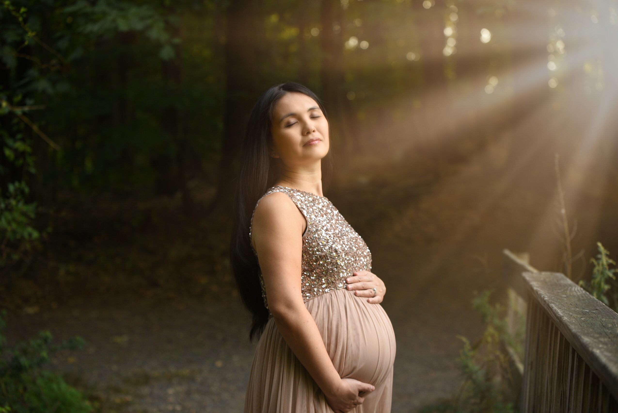 Pregnant person standing beside a wooden railing as sunlight filters through trees.