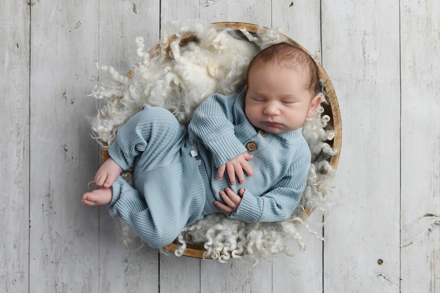 A sleeping baby in a blue outfit lies in a round basket lined with soft fabric on a wooden floor.