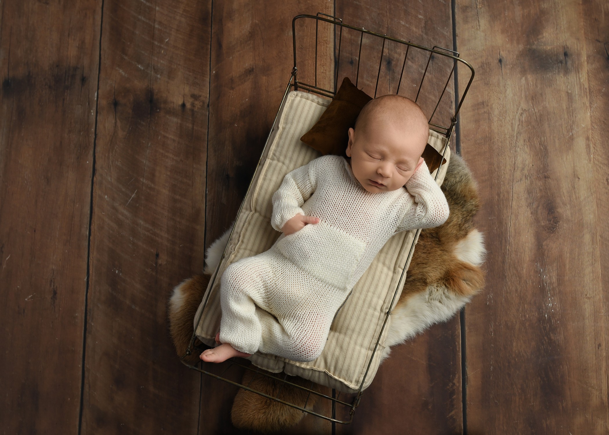 A baby in white clothing lies in a small wire basket on a wooden floor, partly atop a furry blanket.