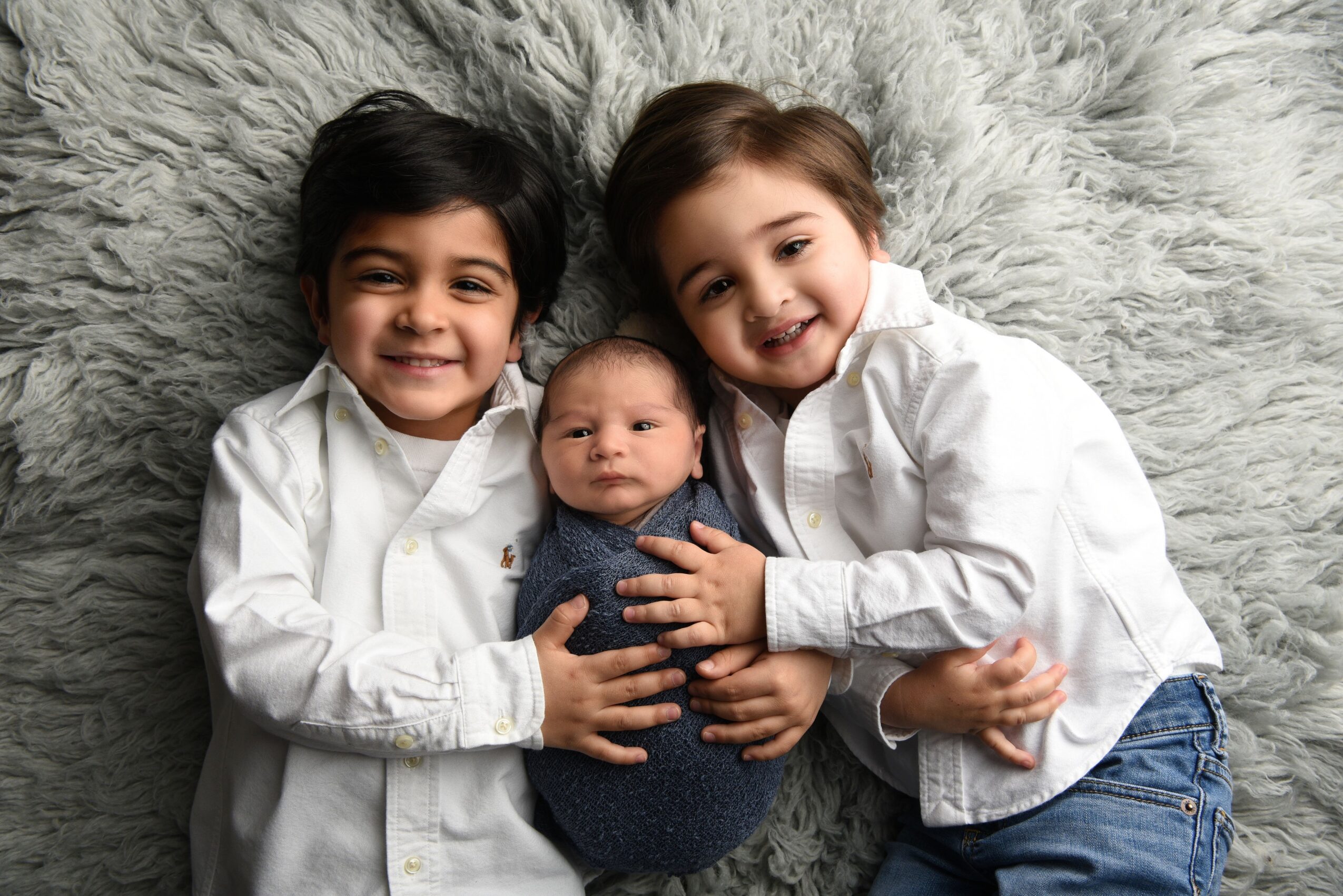 Two children lie beside an infant on a fluffy gray rug.