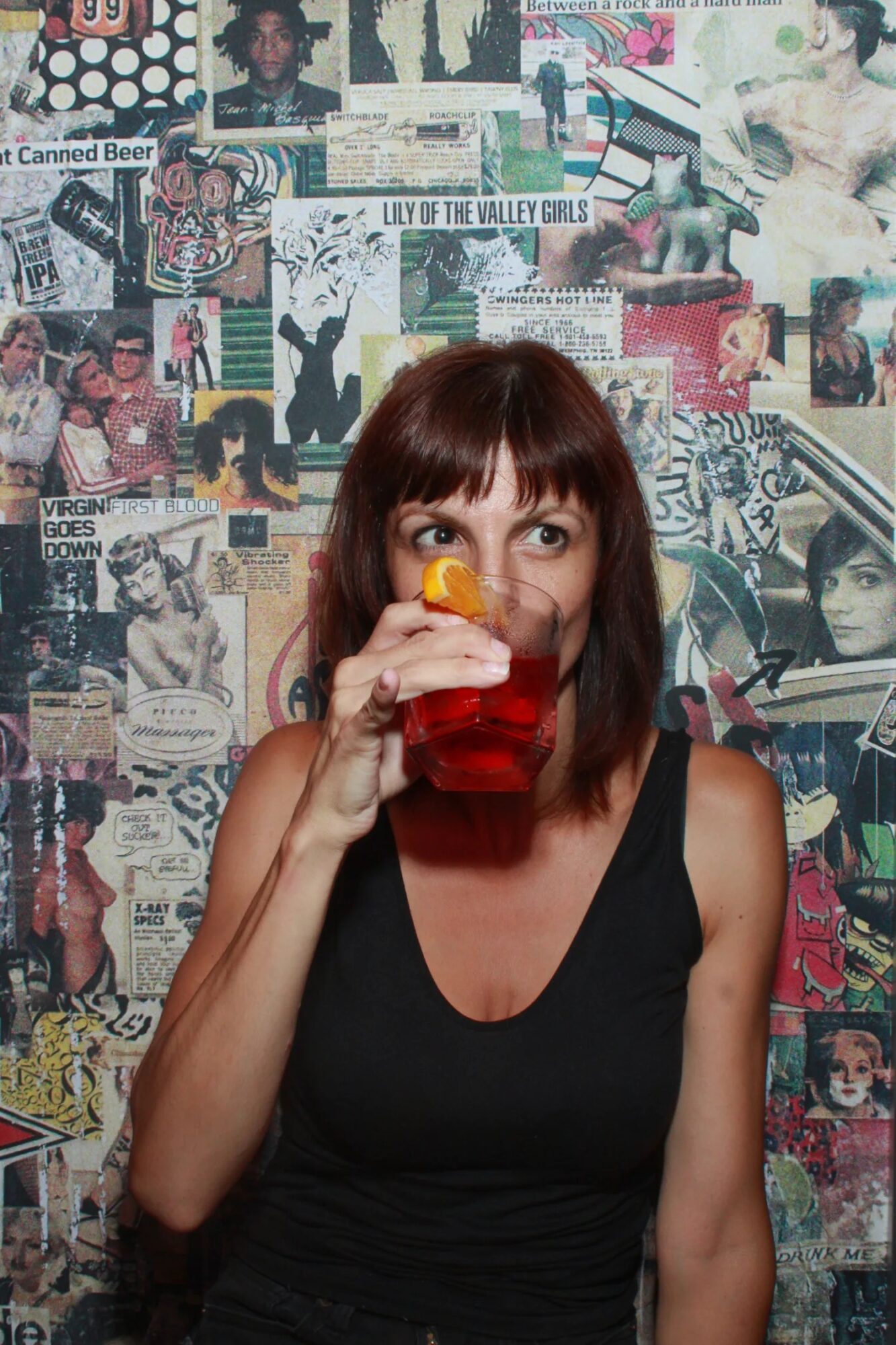 A seated person holds a glass of red liquid near their face, against a wall covered with posters and magazine pages.