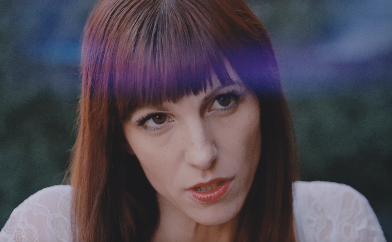 Close-up portrait of a person with straight dark hair and bangs, wearing a white top against a blurred background.