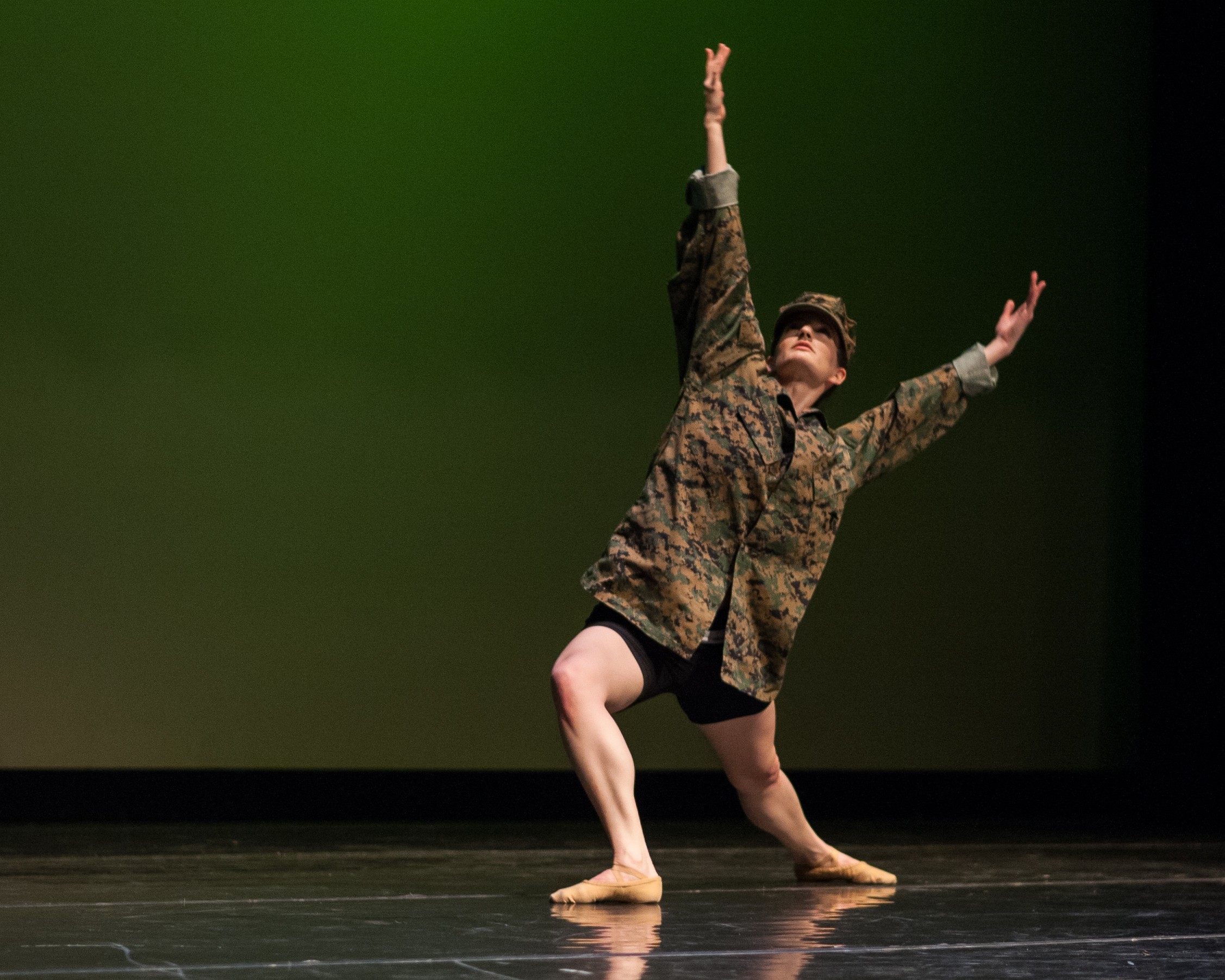 A dancer in a patterned coat balances on one leg with both arms raised on a stage.