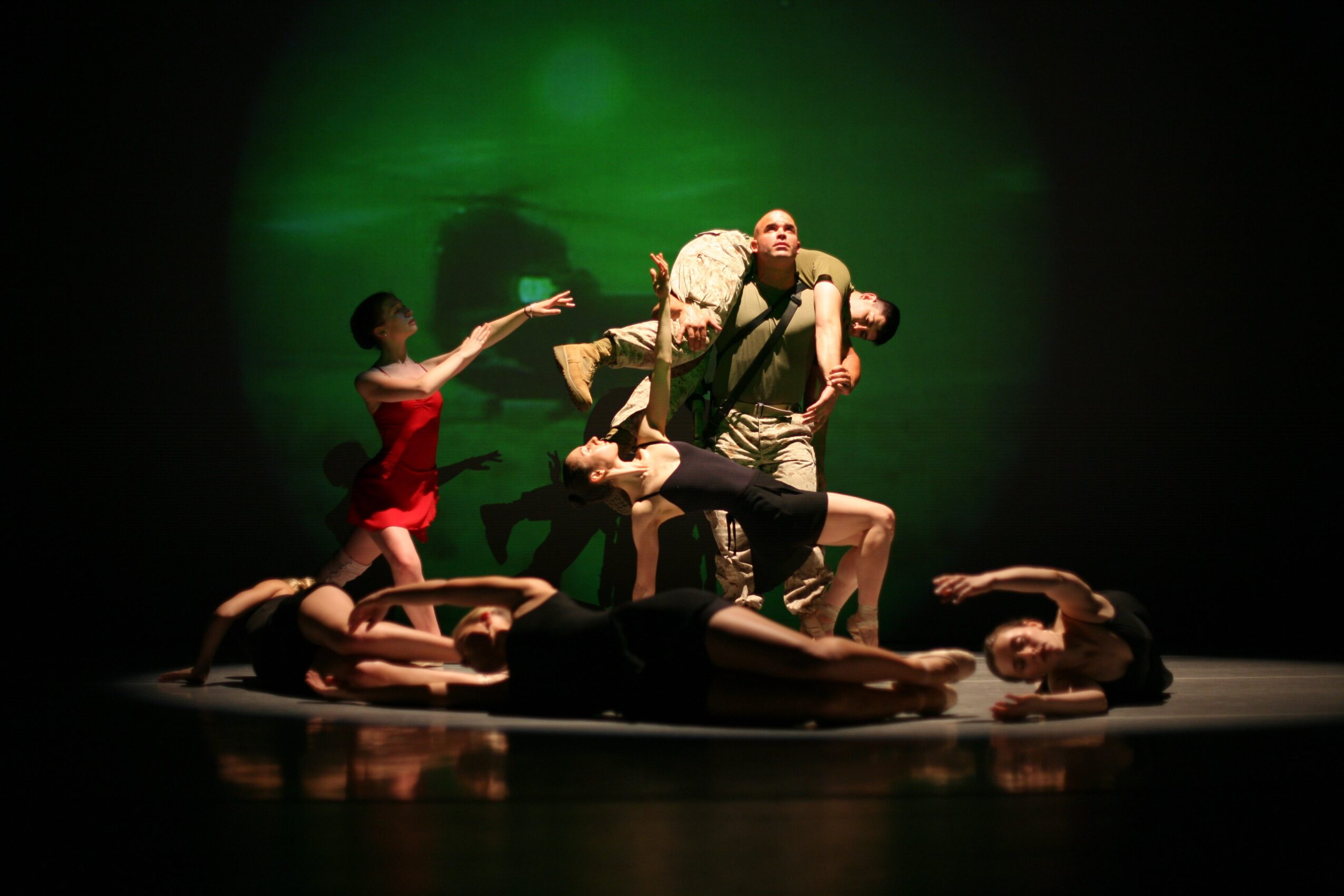 A group of performers forms a layered pose on a stage beneath green lighting.