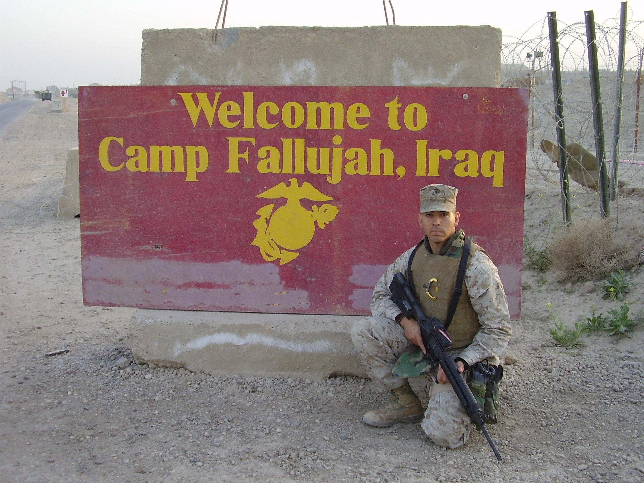 A person holding a rifle kneels beside a sign reading “Welcome to Camp Fallujah, Iraq.”