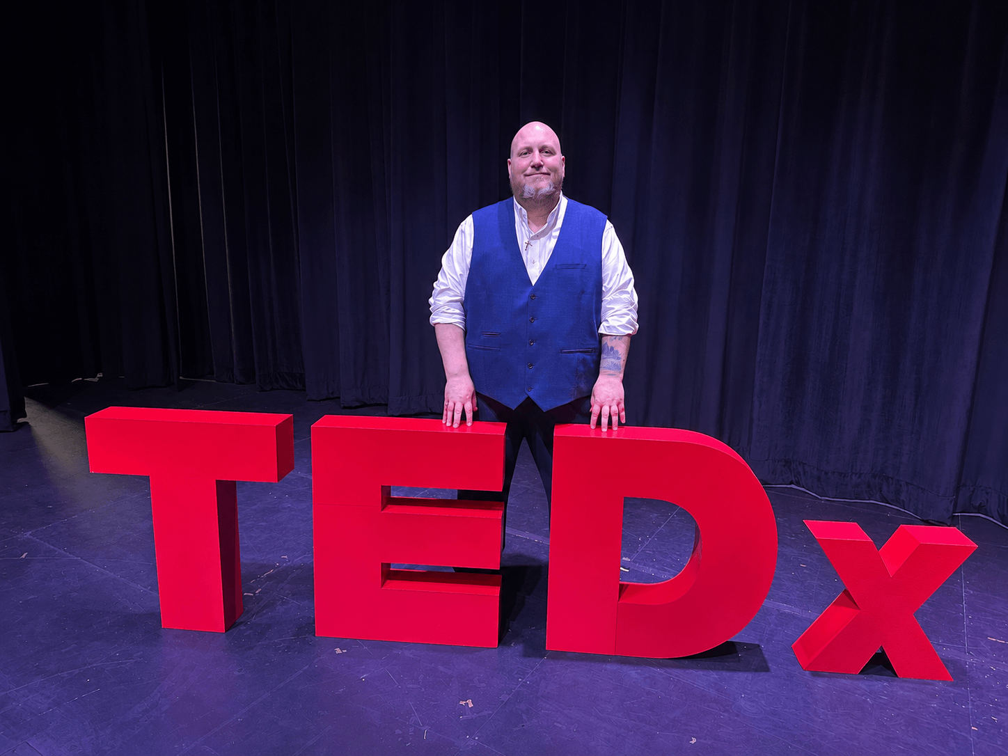 A person stands behind large red TEDx letters on a stage with dark curtains.