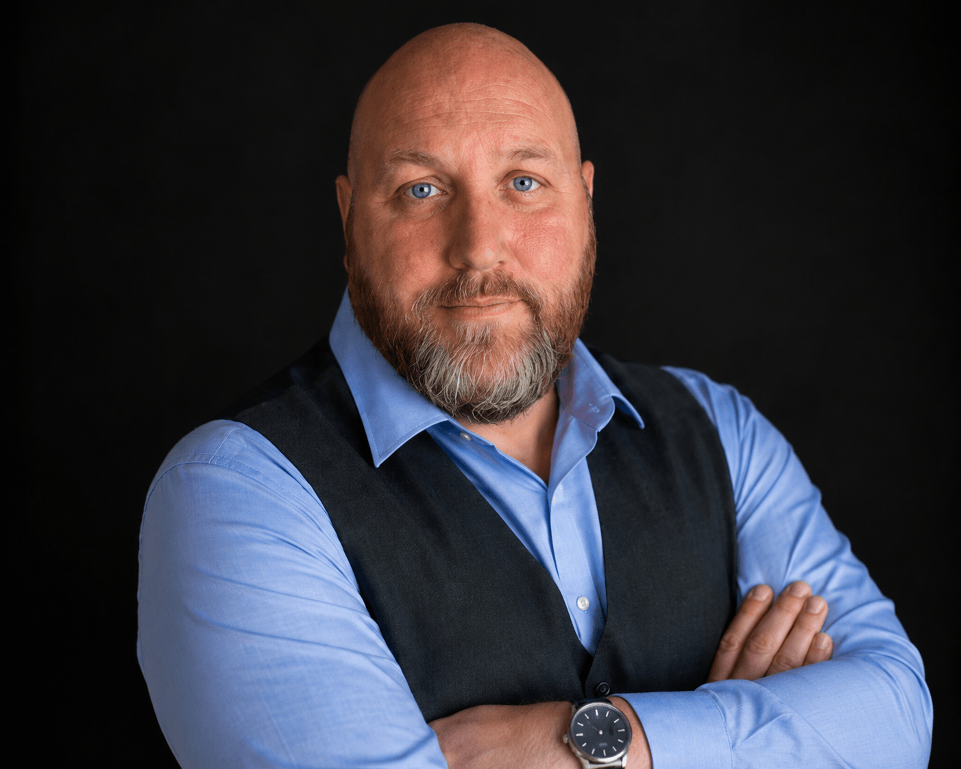 Bald bearded person in a blue shirt and dark vest, arms crossed against a black background.