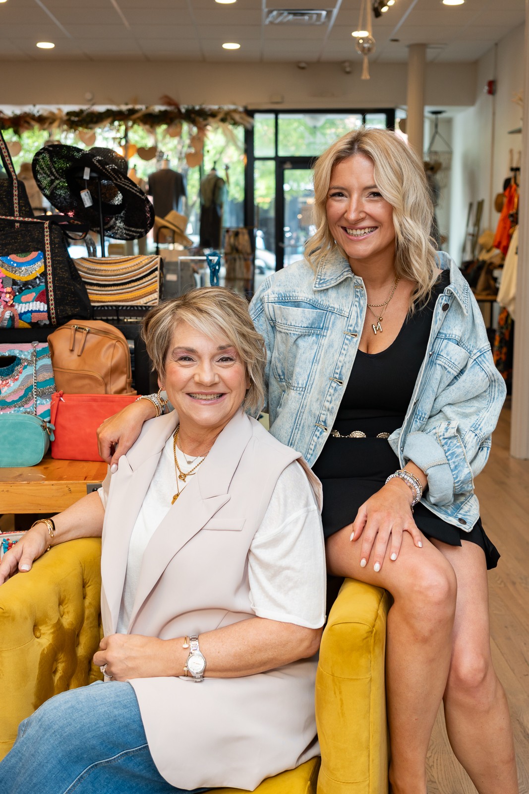 Two people pose together in a boutique, one seated in a yellow chair and the other standing beside her.