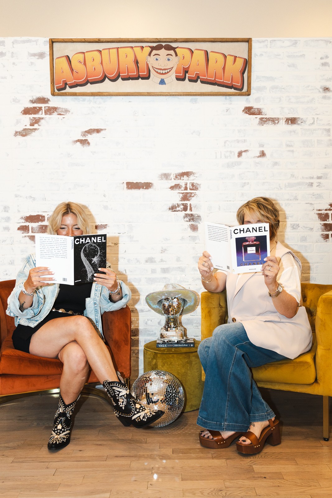 Two people sit in chairs holding Chanel products beneath an Asbury Park sign.