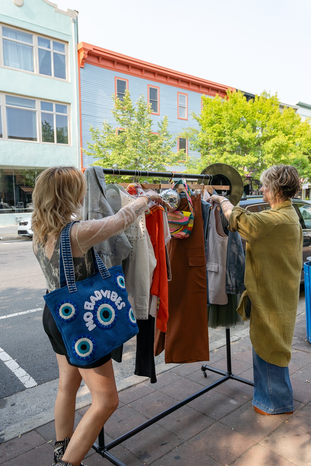 Two people browse clothing on a sidewalk rack beside a blue building and leafy trees.