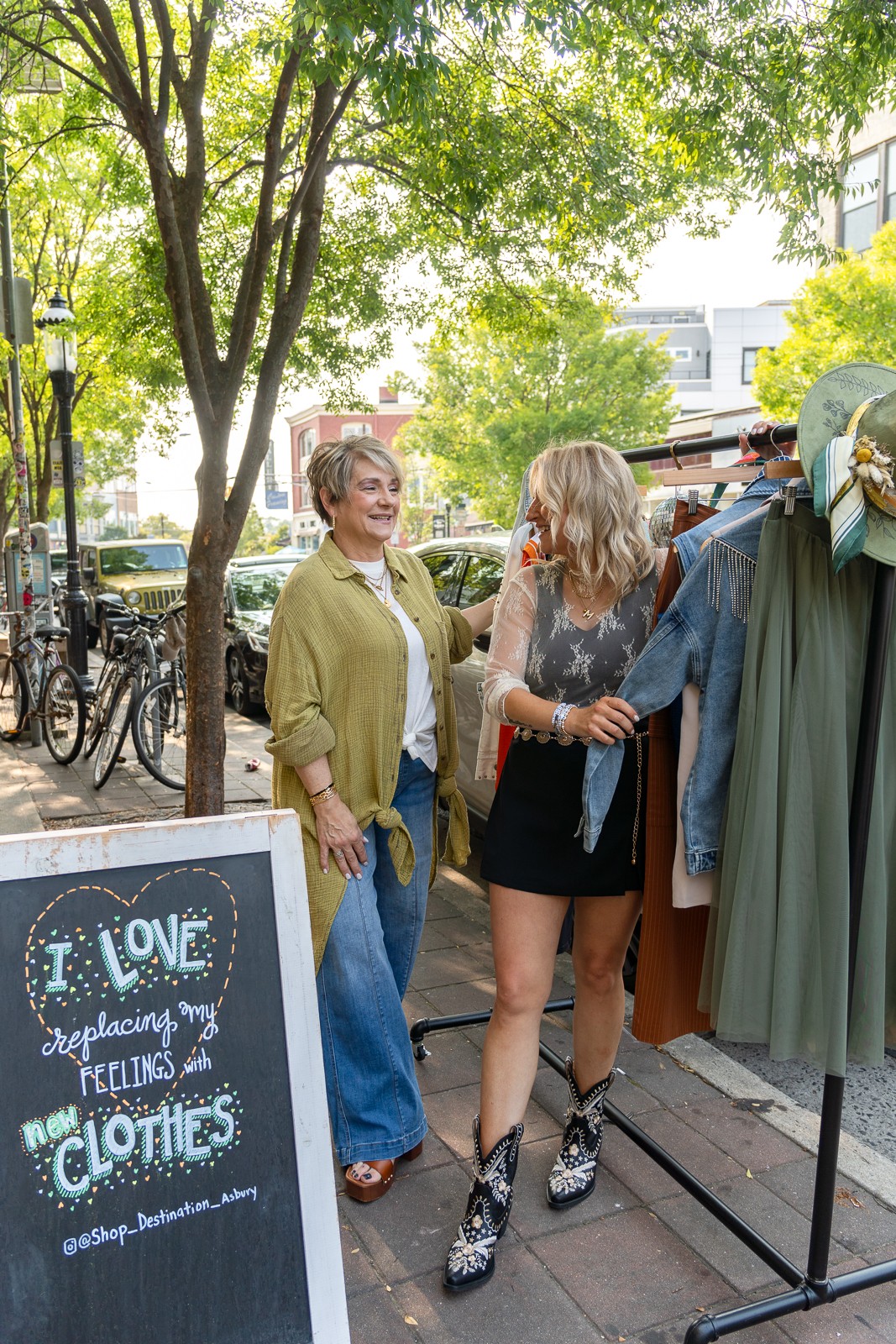 Two people stand beside a clothing rack and chalkboard sign on a tree-lined sidewalk.