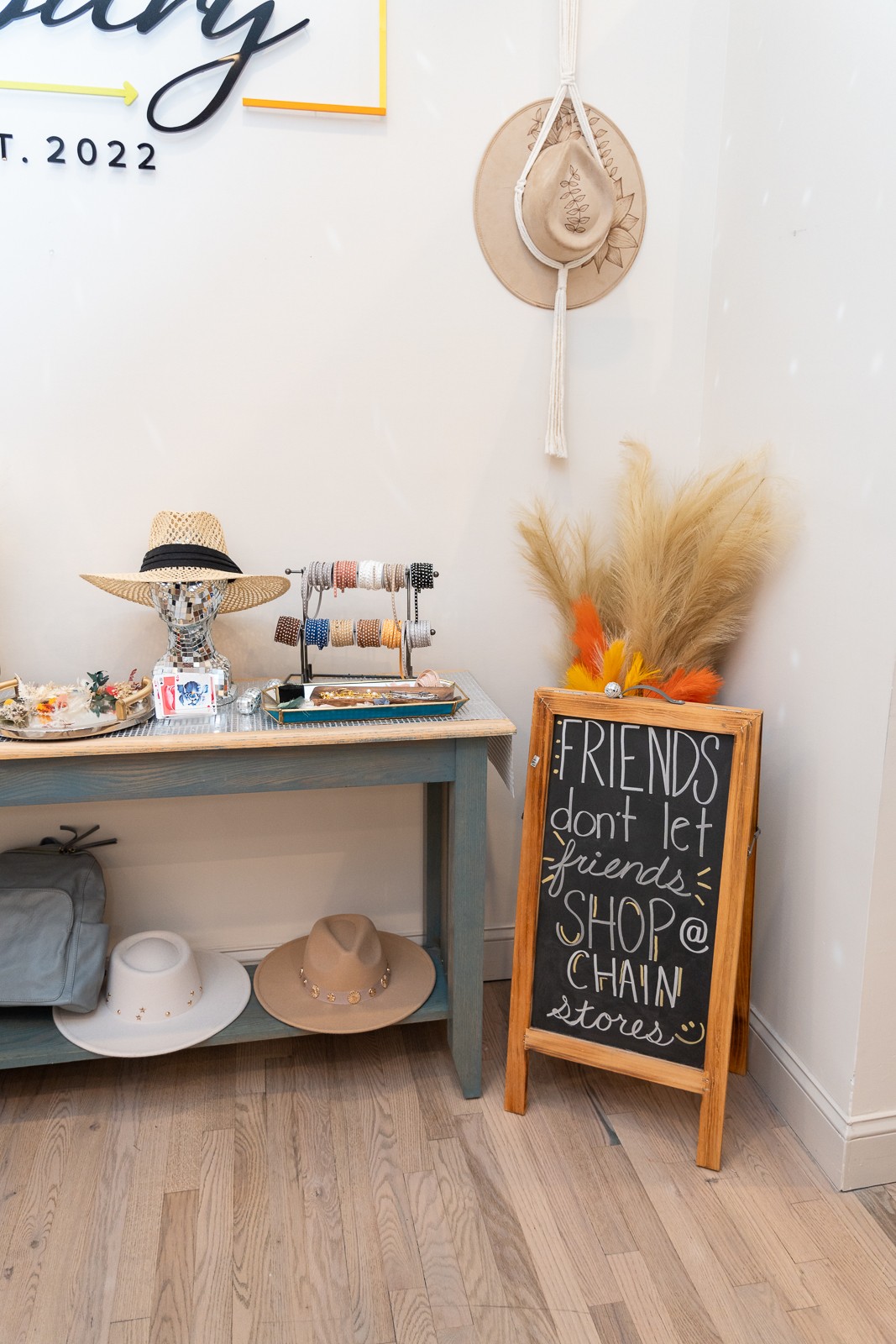 Boutique corner with a display table, hats, dried grasses, and a chalkboard sign reading “FRIENDS don’t let friends SHOP alone.”