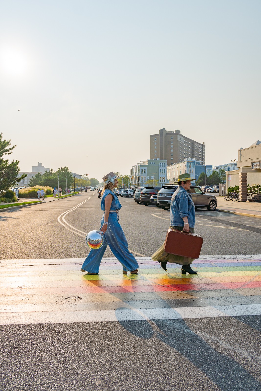 Two people cross a rainbow-colored pedestrian crossing on a sunlit urban street.