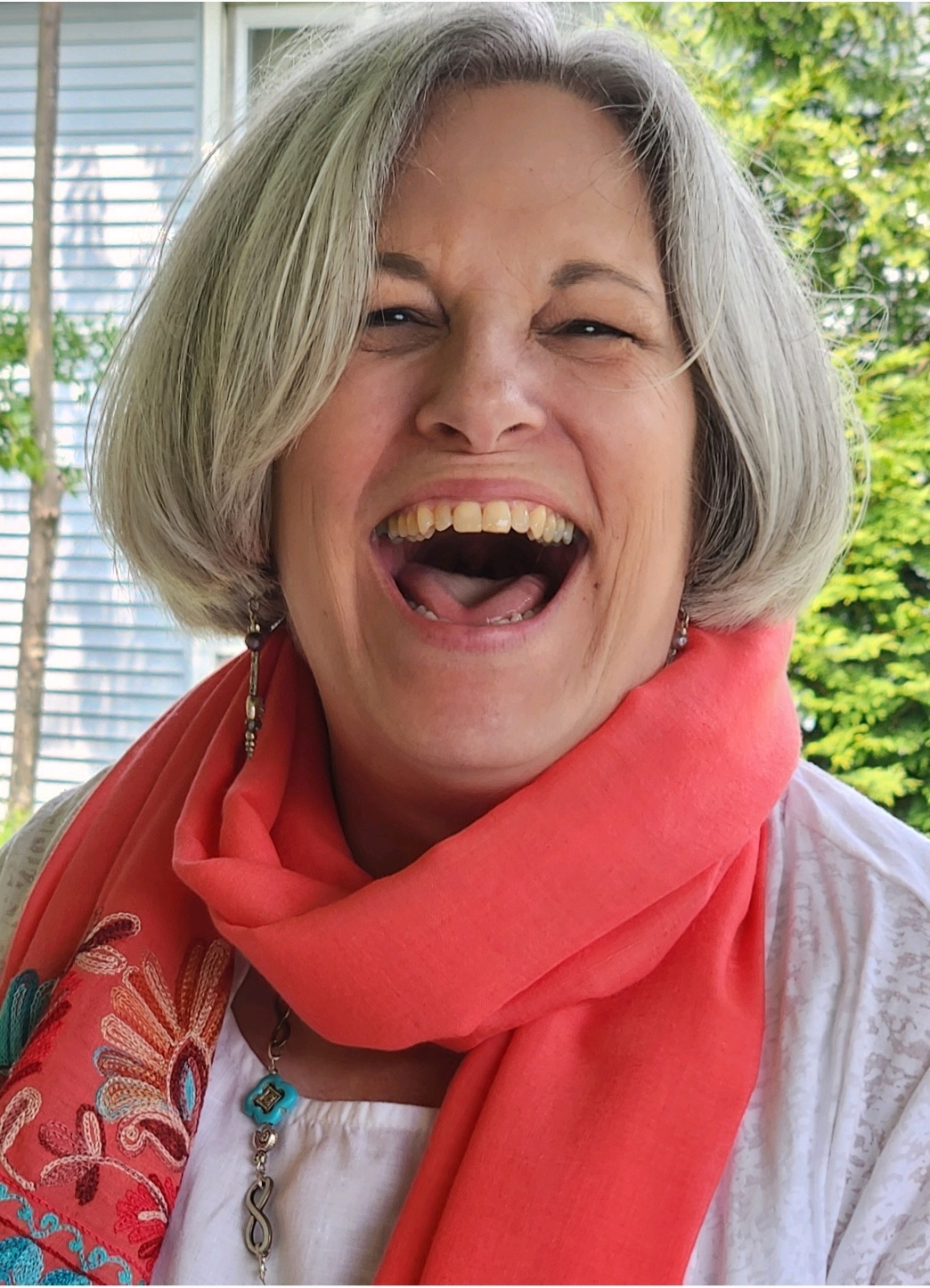 A person with short gray hair wearing a bright coral scarf, smiling in front of a window and greenery.
