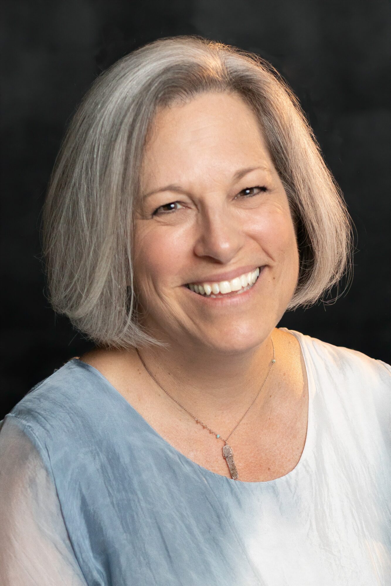 Portrait of a smiling person with a short gray bob, wearing a blue and white top against a dark background.
