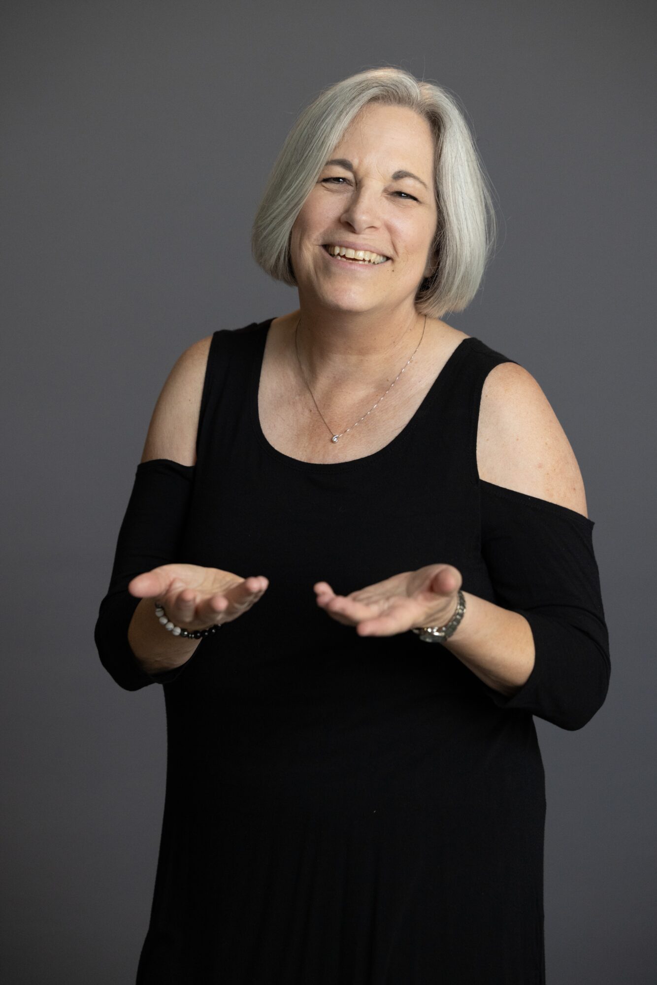 A person with short gray hair wearing a black dress holds both hands out against a gray background.