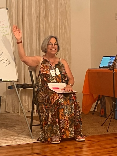 A seated person raises one hand beside a whiteboard in a room with curtains and a table.